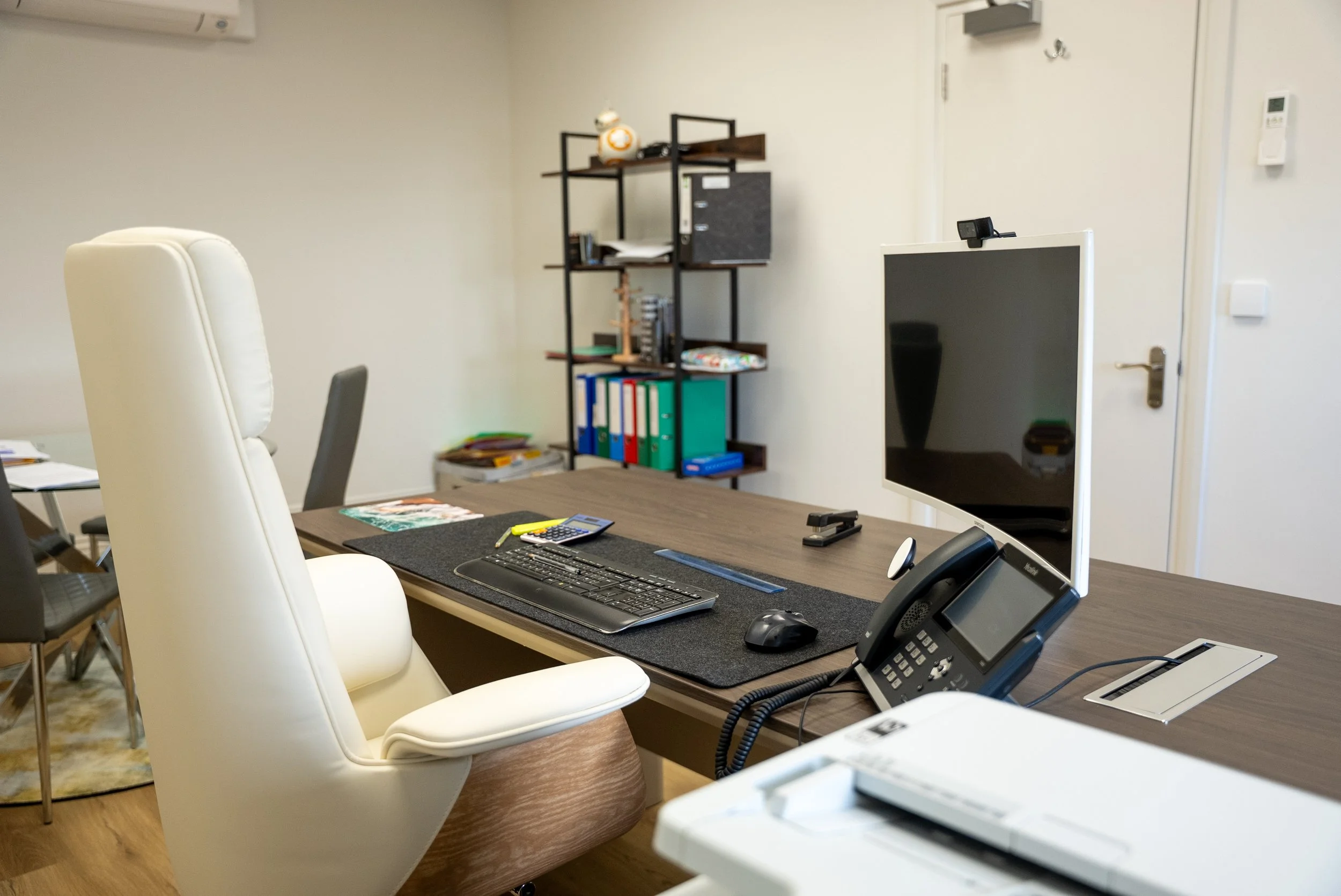 An office desk with a white high-back chair, computer monitor, keyboard, mouse, phone, calculator, and documents, with a bookshelf in the background.