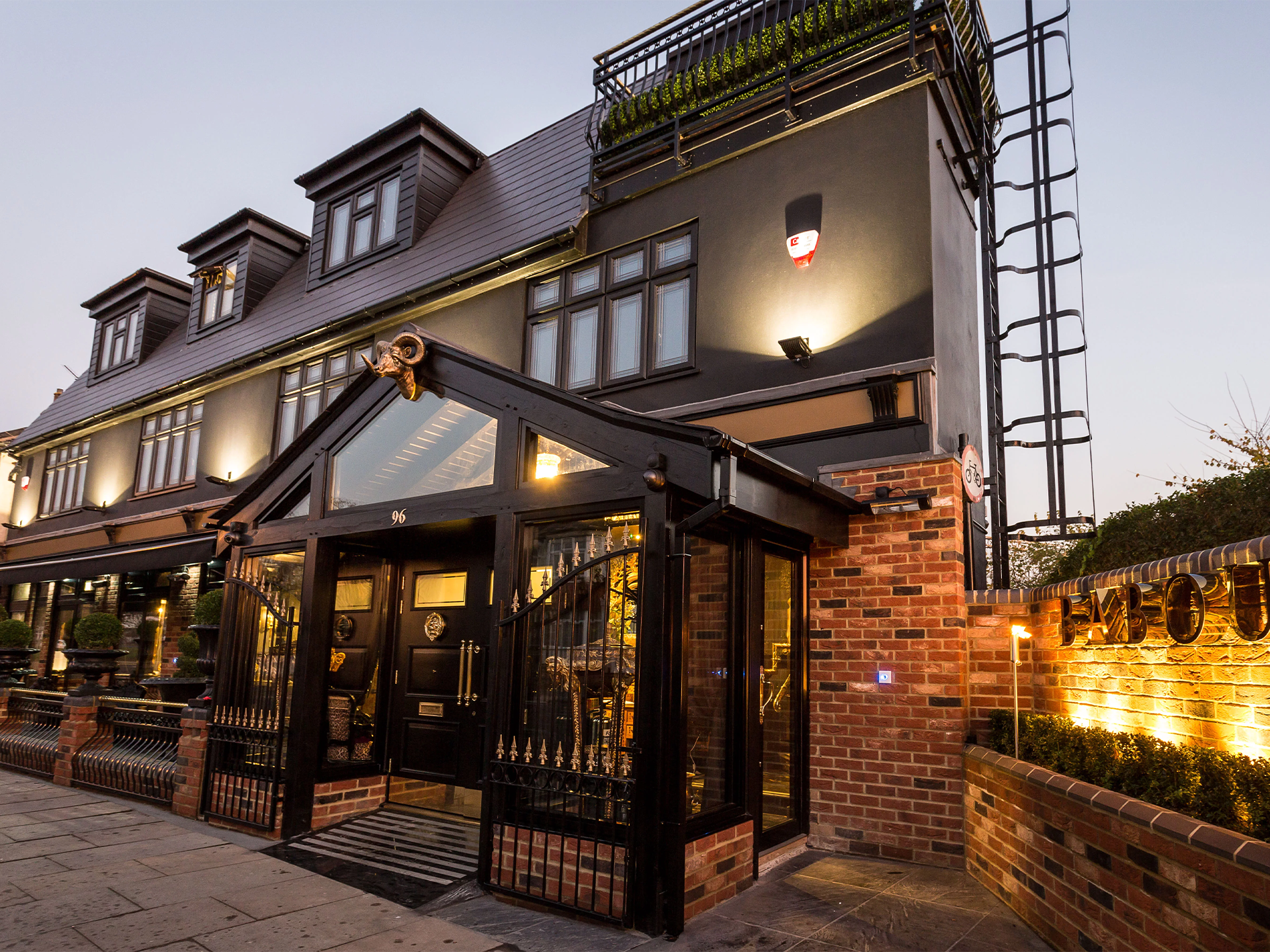 Exterior view of a multi-story building with black and brick façade, decorative lantern, and metal gate, illuminated at dusk, with a sign reading "Baboush" on the brick wall.