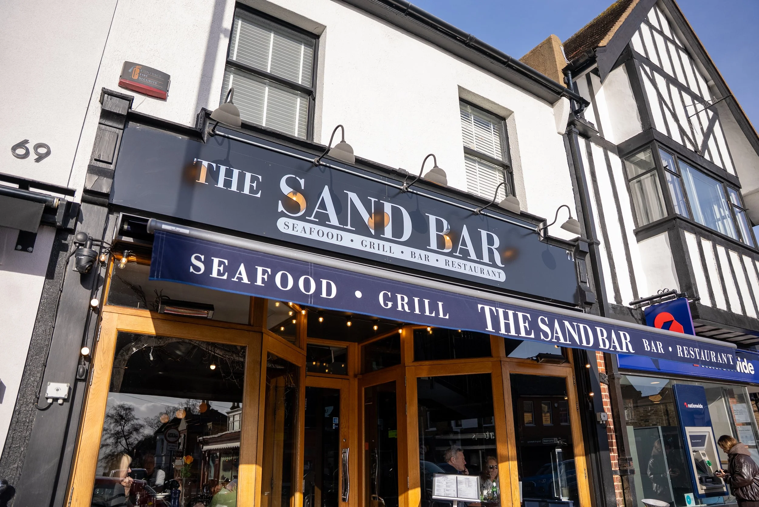 Exterior view of The Sand Bar restaurant with signage indicating it serves seafood, grill, bar. The restaurant has wooden framed glass doors, and several people are inside. There is a blue awning with the restaurant's name repeated, and the building features white walls with black timber framing.