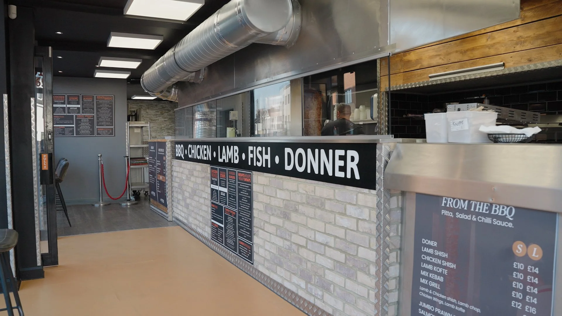 Inside a fast-food restaurant counter with a black sign listing BBQ, chicken, lamb, fish, and dinner options, with menus and prices displayed on the wall and counter.