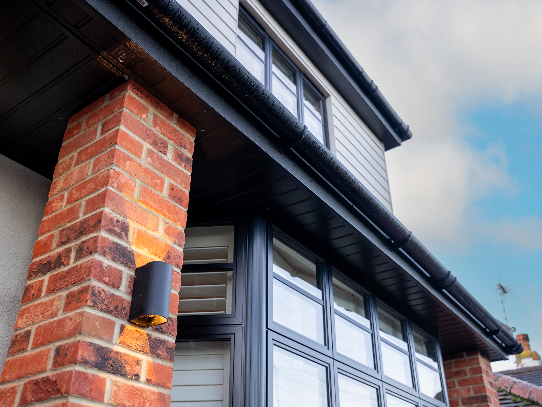Close-up of a modern house corner with brick columns, black metal window frames, and exterior wall siding, under a partly cloudy sky.