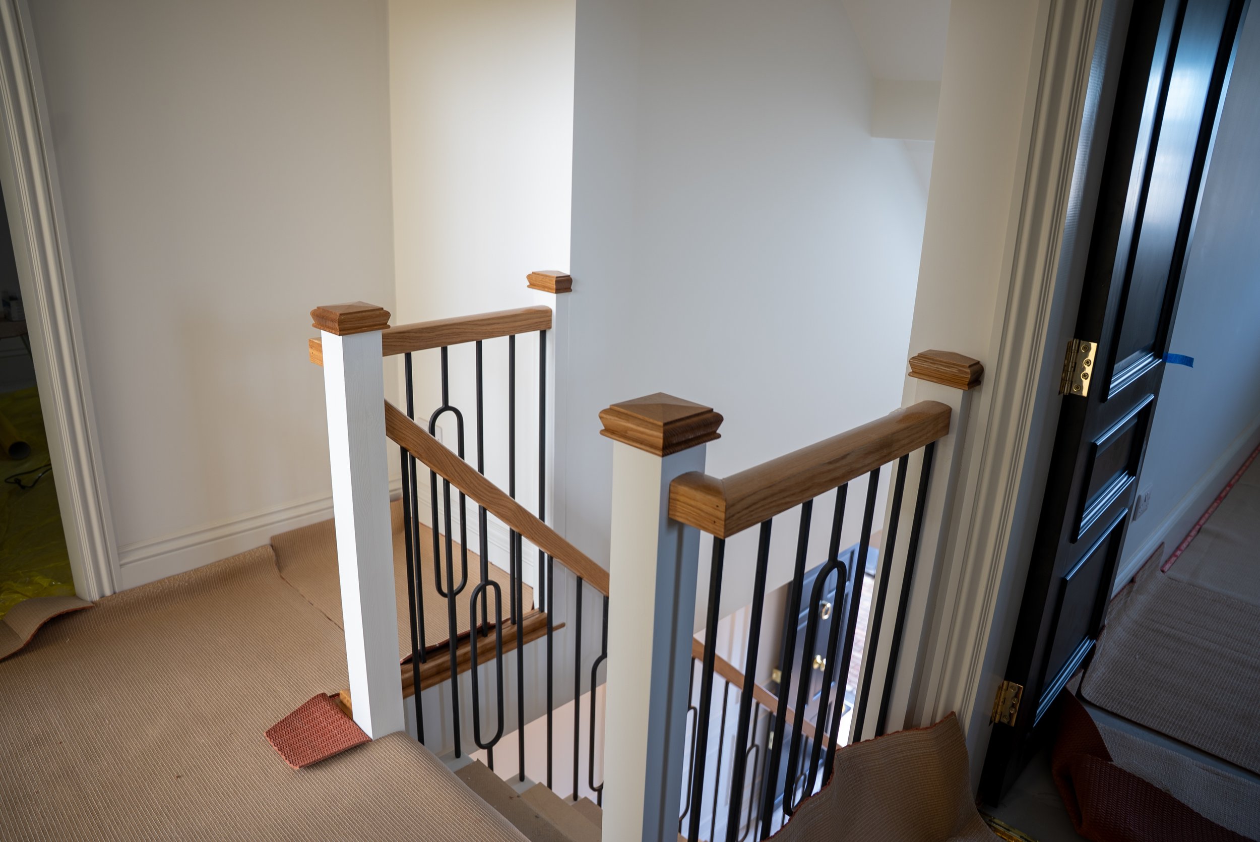 Interior view of a staircase with wooden handrails and black metal balusters, seen from the top floor looking down.