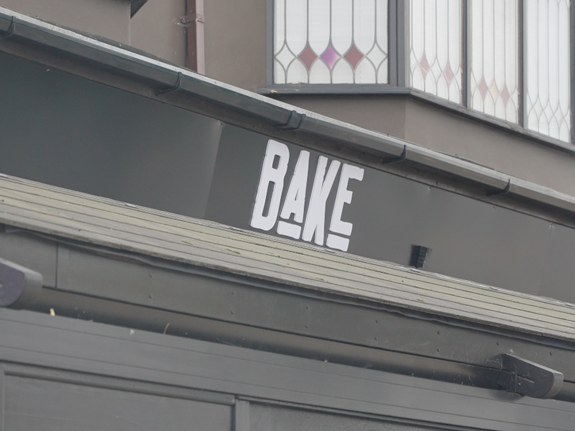 Close-up of a building with a sign reading 'BAKE' in large white letters on a dark background, with a window featuring decorative stained glass panels above.