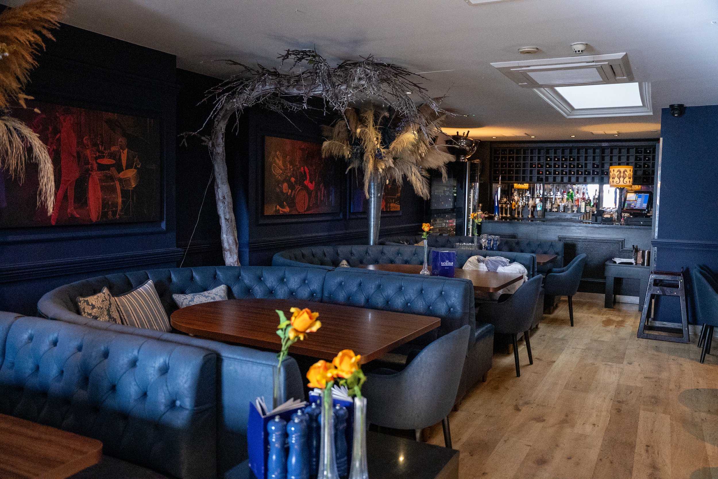 Interior of a restaurant or bar with blue tufted booths, wooden tables, and floral arrangements in vases. A large decorative tree with branches extends from the center of the seating area. The bar area with bottles and glasses is visible in the background.