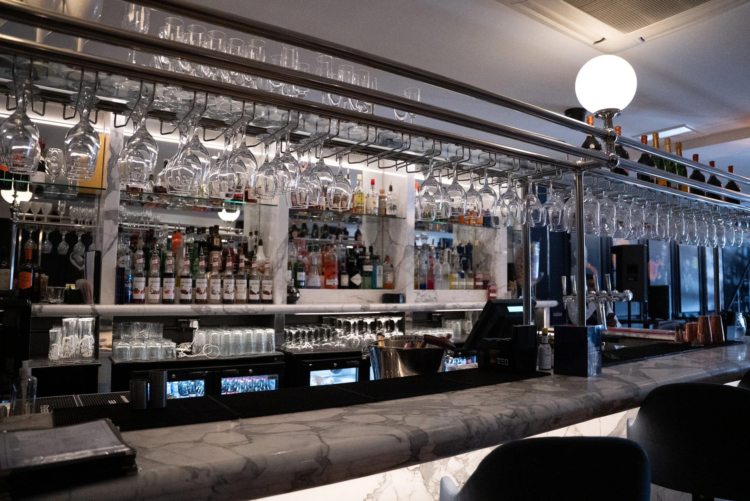 Empty bar with hanging glasses, liquor bottles, and a marble countertop in a dimly lit lounge or restaurant.