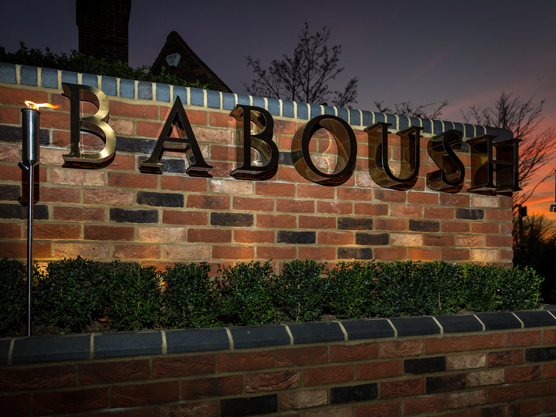 A brick wall with the word 'BABOUSH' displayed in large black 3D letters. There is a small hedge in front of the wall, and two lights on either side illuminating the sign. The sky is at sunset with a few trees visible in the background.