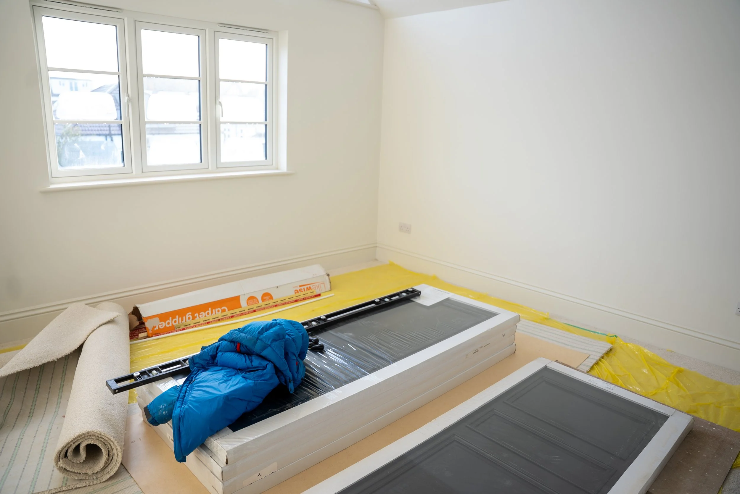 Room under renovation with a large window, yellow plastic sheets on the floor, a door, a coat, and construction materials like a level and a door panel.