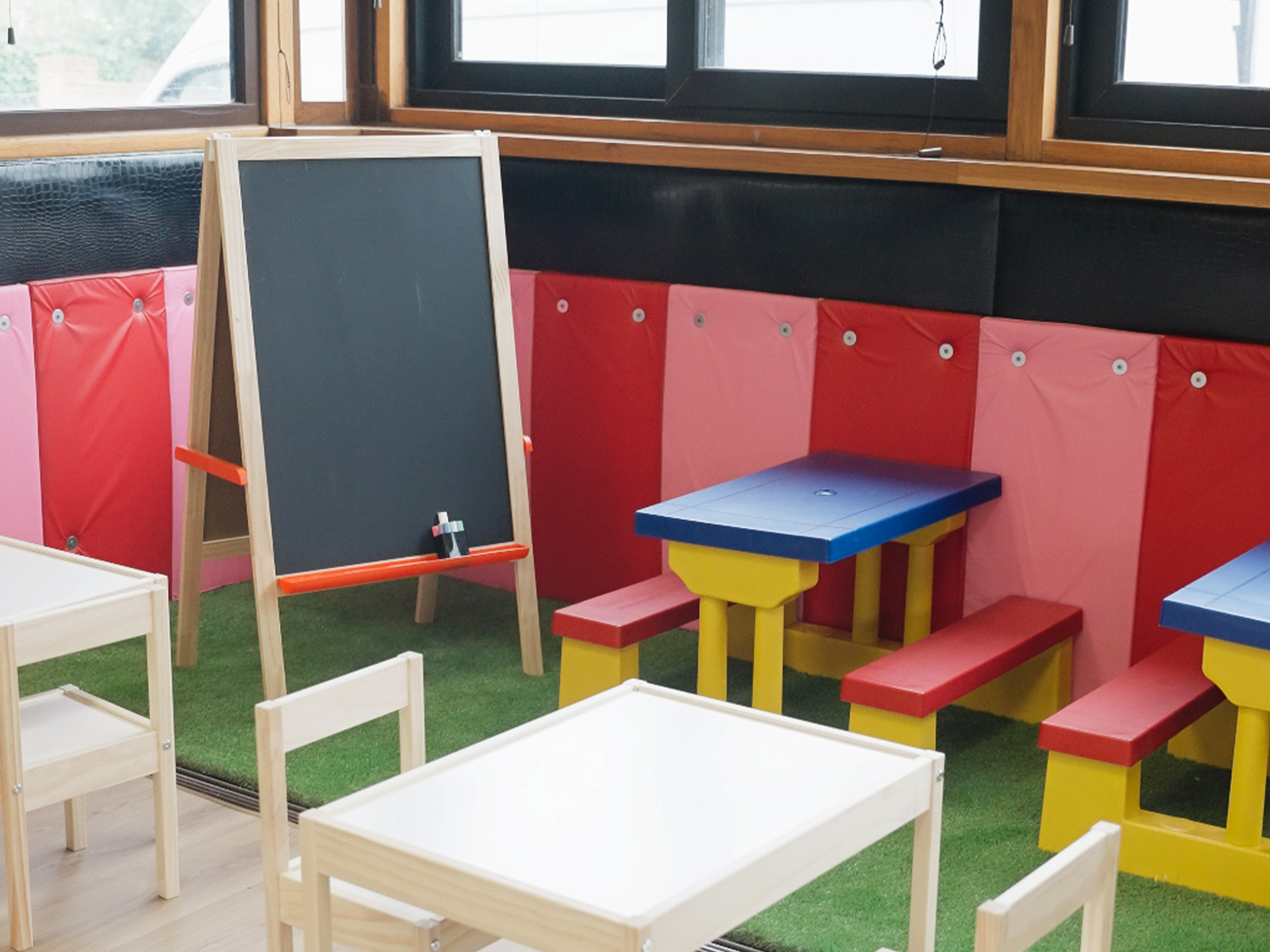 Colourful children's play area with small wooden tables and chairs, a chalkboard, and a padded corner with a small table and benches, inside a room by windows.