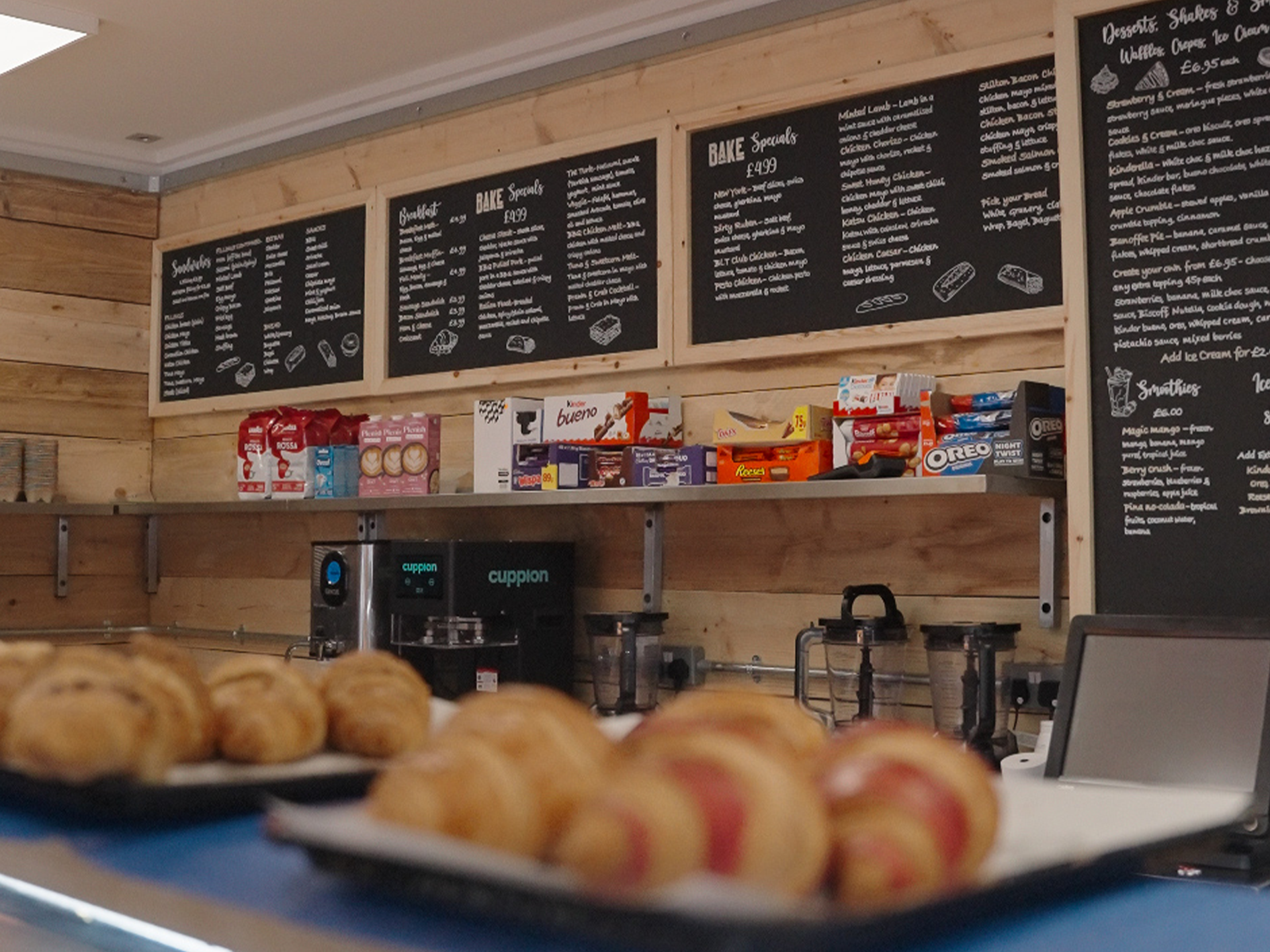Close-up of baked goods on trays in a cafe or bakery, with a wooden wall and menu boards in the background.
