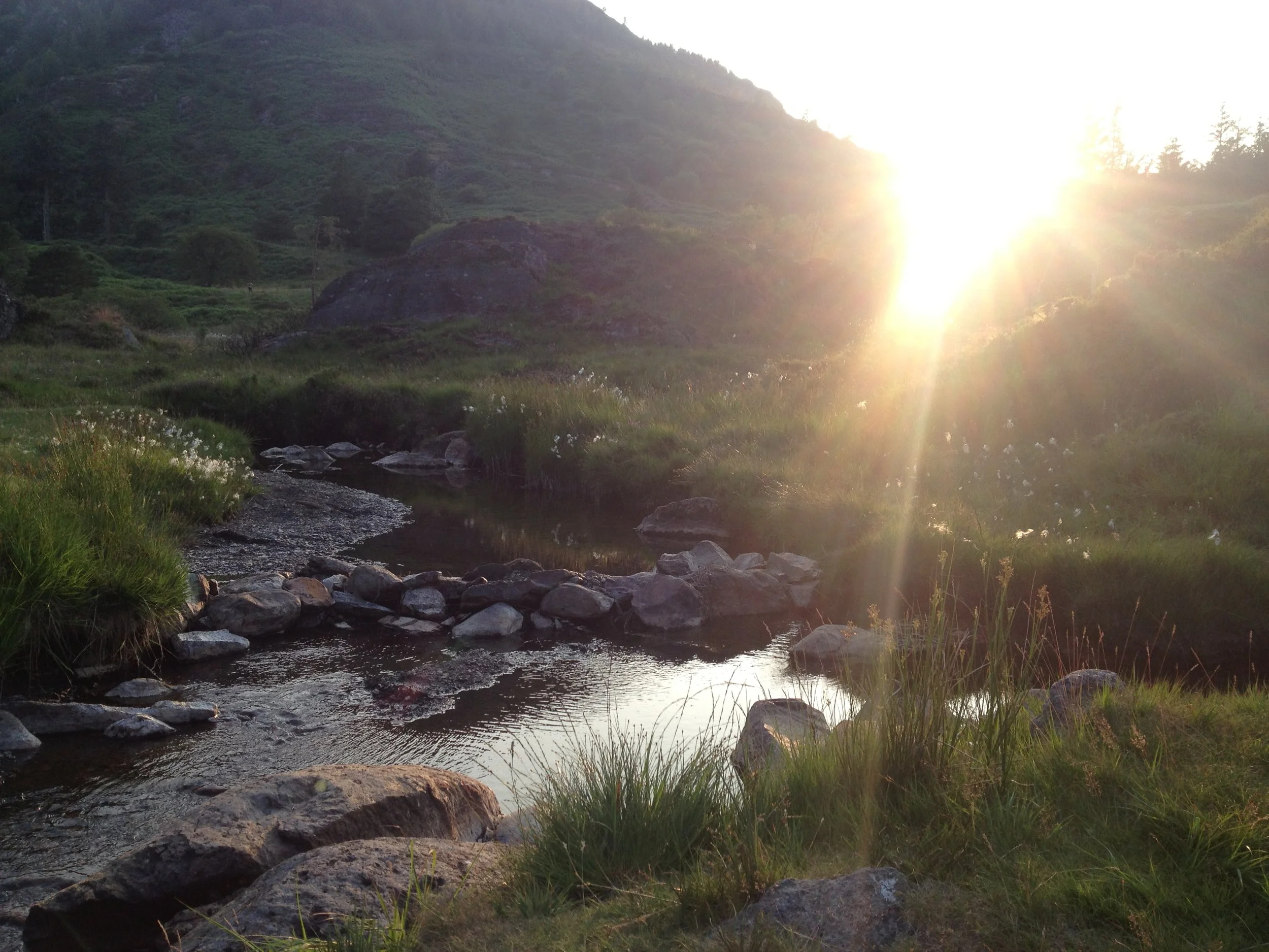 Sunset over a small river flowing through a grassy, rocky landscape with green hills in the background.