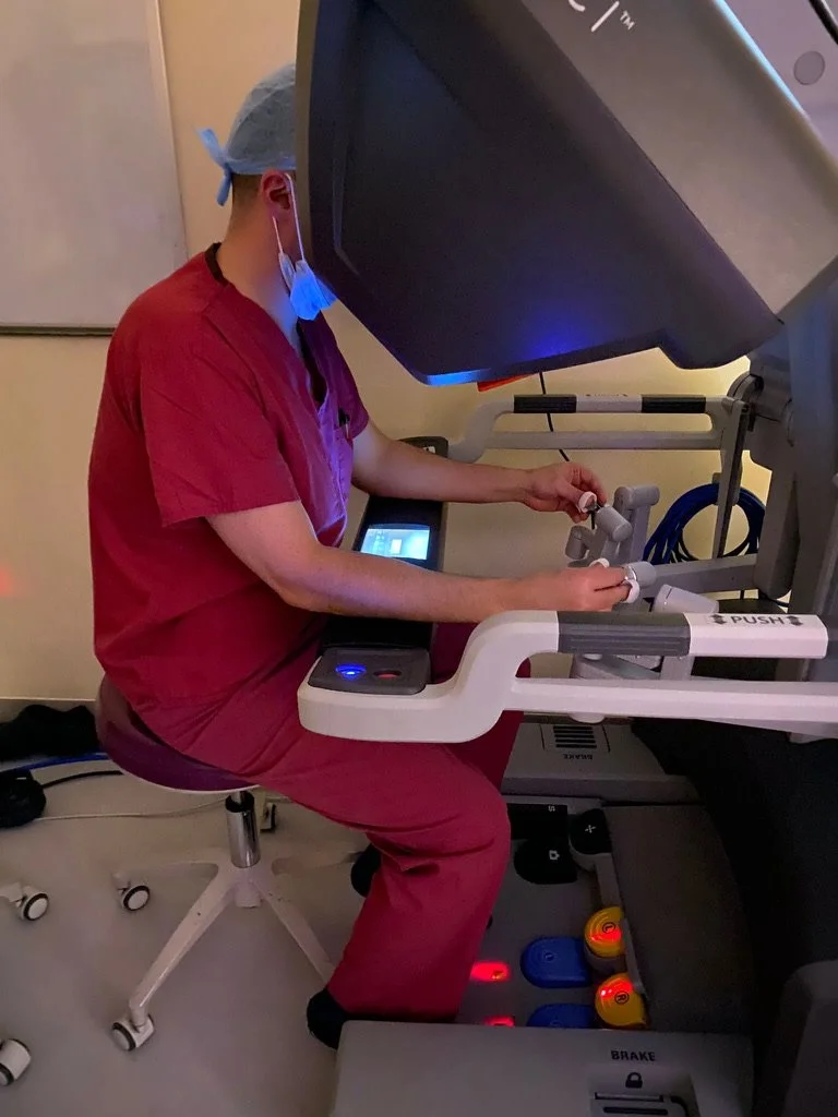 A medical professional in red scrubs operating a robotic surgical system in a hospital or clinical setting.
