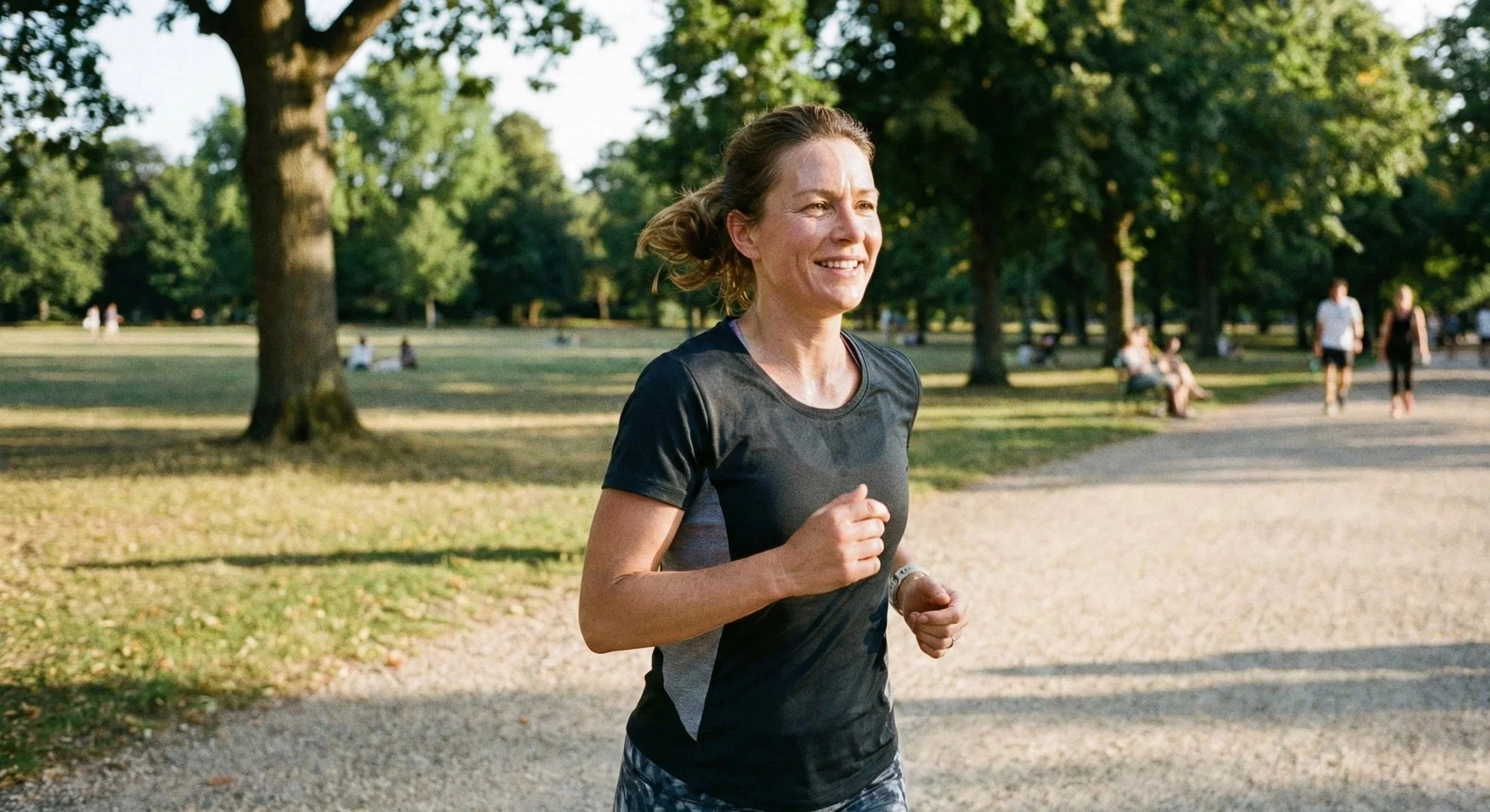 A woman jogging in a park on a sunny day, smiling and wearing a black athletic shirt and patterned workout pants.