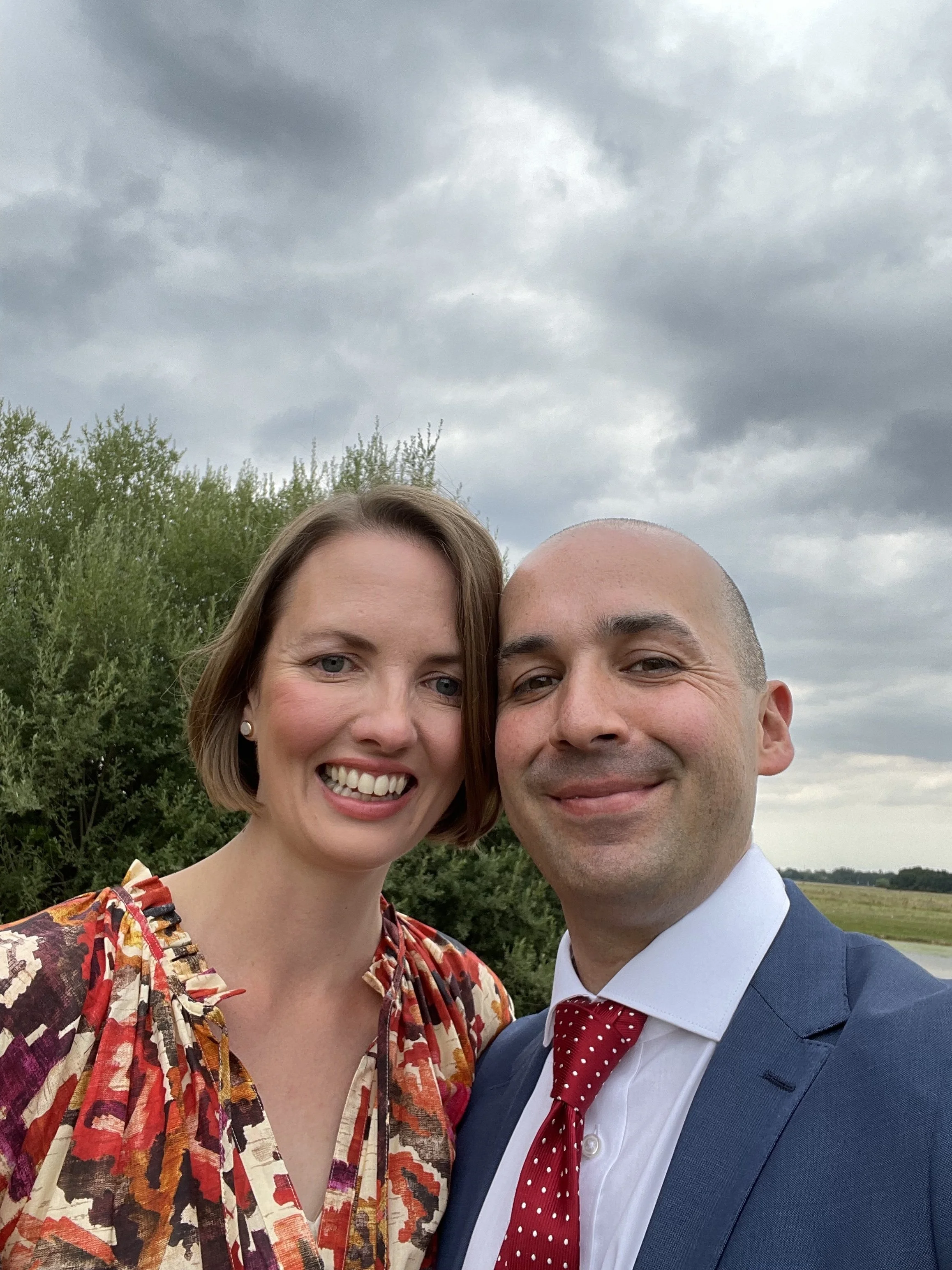 A smiling woman with short brown hair wearing a colorful, patterned blouse and pearl earrings, standing next to a smiling man with a shaved head wearing a navy suit, white shirt, and red polka dot tie, outdoors with greenery and cloudy sky in the background.