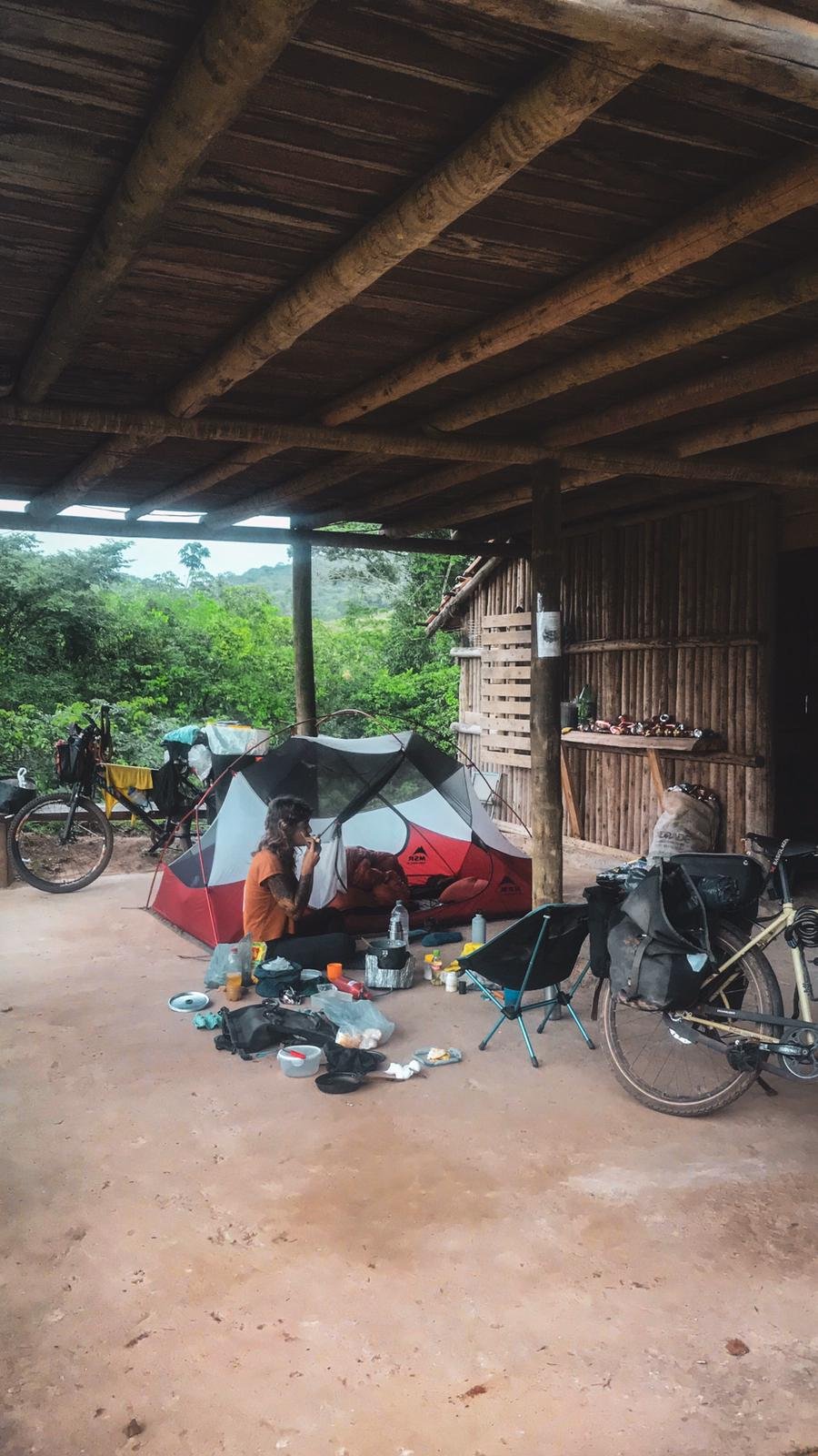 Person sitzt vor einem Zelt im Freien unter einem Holzdach, umgeben von Fahrrädern und Campingausrüstung, in einer ländlichen, grünen Umgebung.