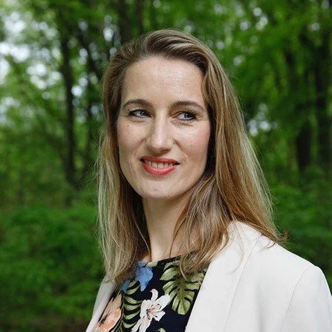 Portret van een vrouw met lang bruin haar, lachend, buiten in een bosrijke omgeving.