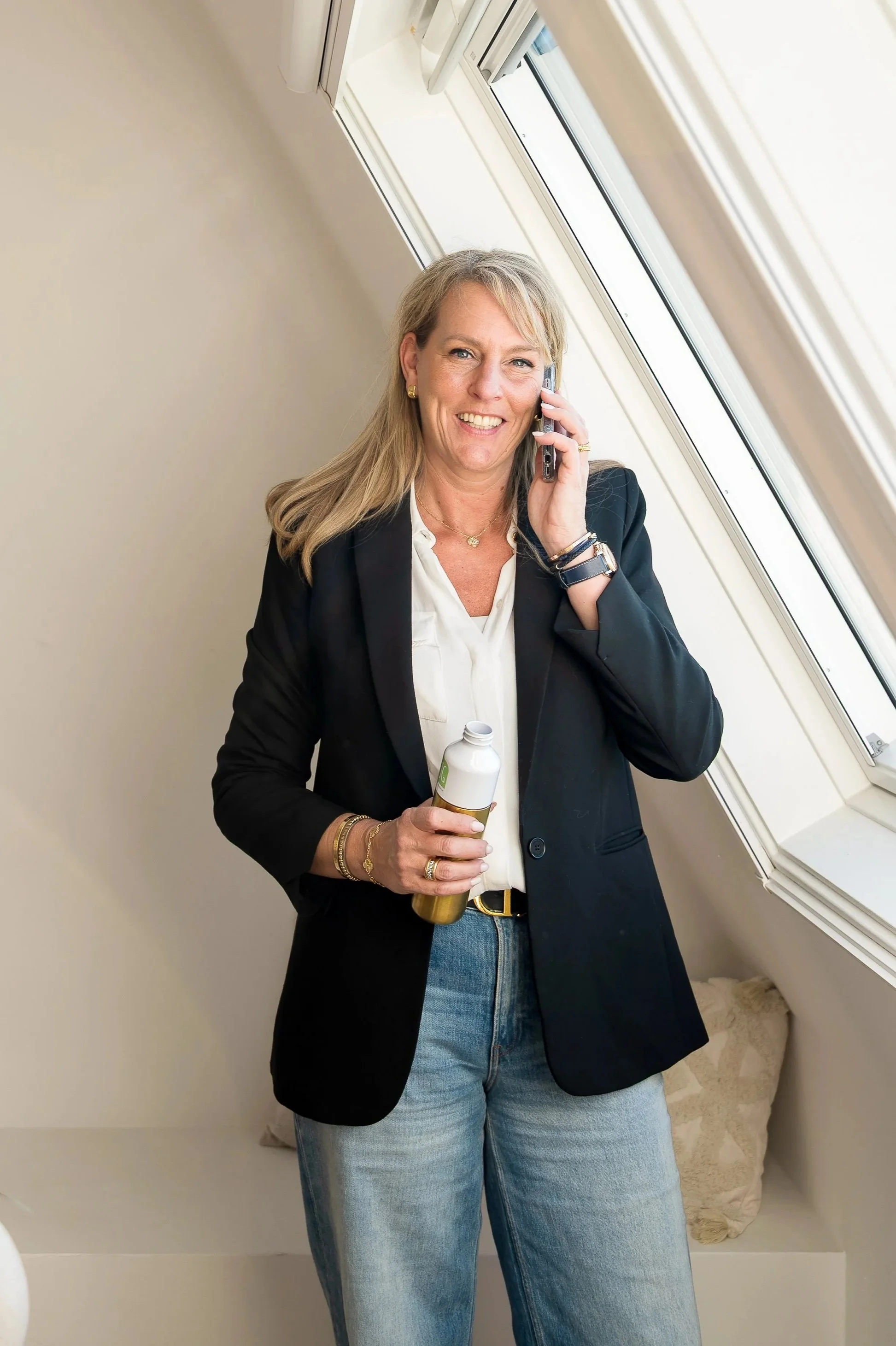 Een glimlachende vrouw met blond haar in een zwart jasje en blauwe spijkerbroek, die spreekt op een mobiele telefoon. Ze houdt een fles drinken in haar rechterhand en staat bij een schuifraam. De ruimte heeft een lichte muur en een zacht tapijt.