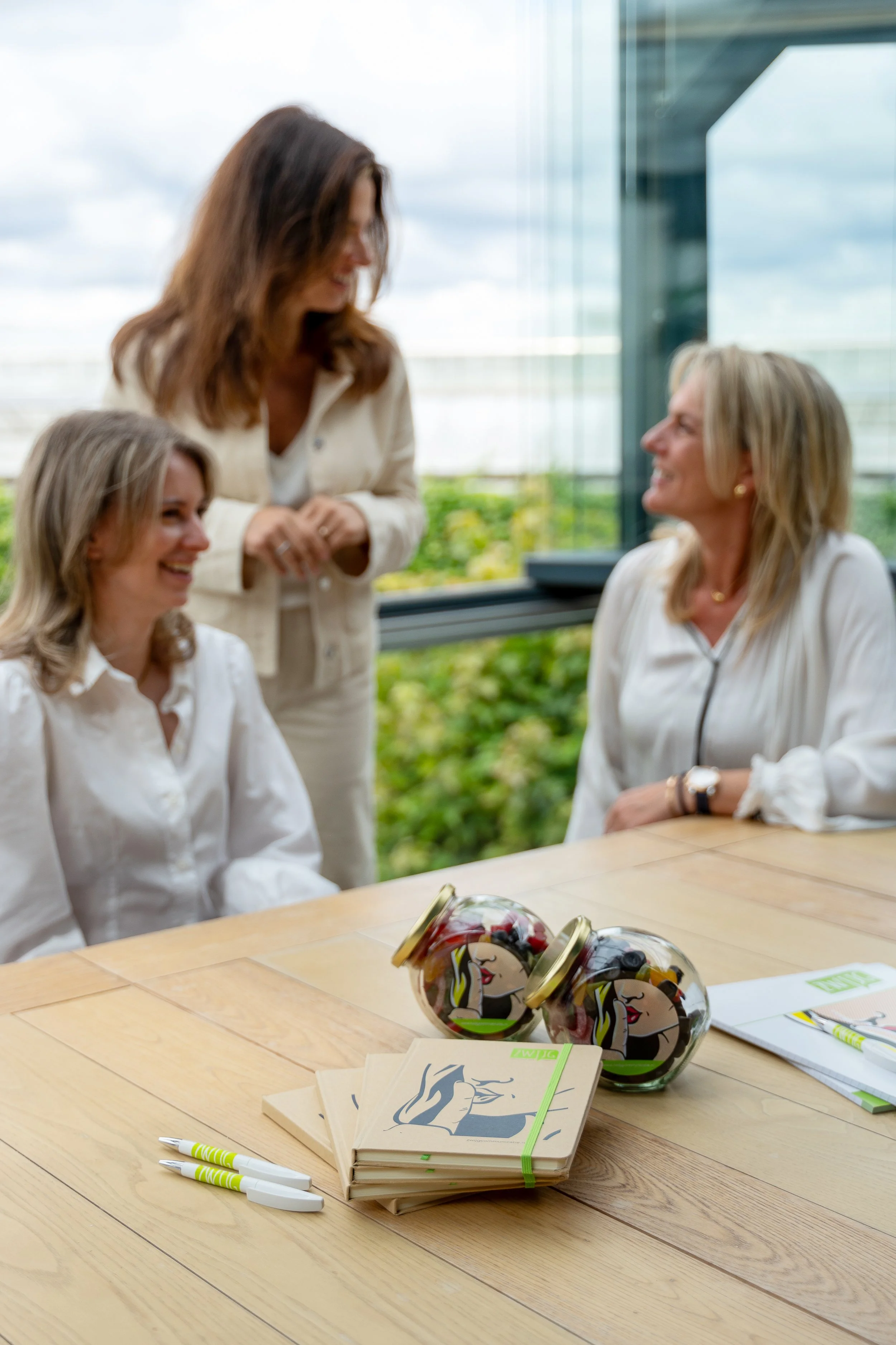 Een tafel met enkele boekjes, pen, en potjes gevuld met fruit, op de achtergrond zitten vier vrouwen die lachen en praten in een groene omgeving.
