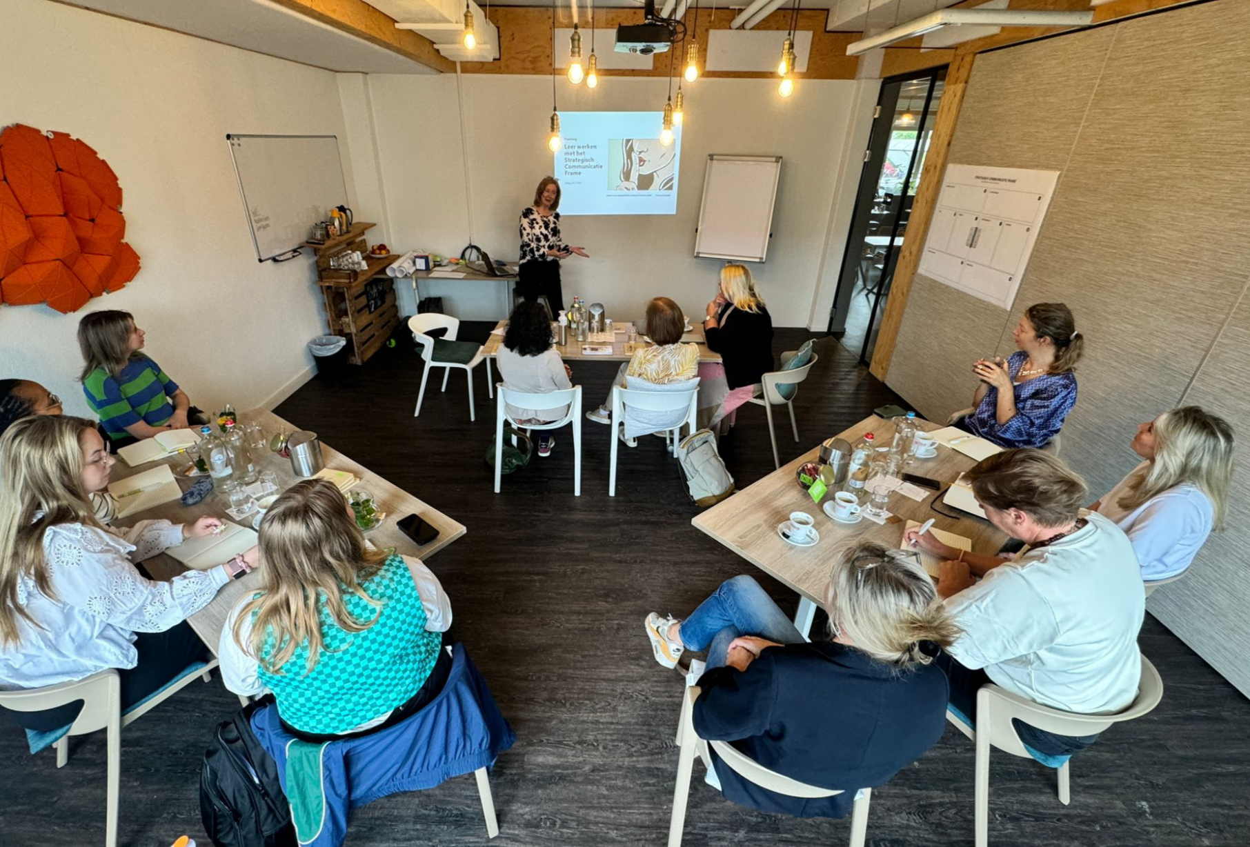 Een groep mensen zit rond tafels in een vergaderruimte terwijl een vrouw voor een scherm spreekt. Op de muur hangen whiteboards en er liggen koffie- en theeservoers op de tafels.