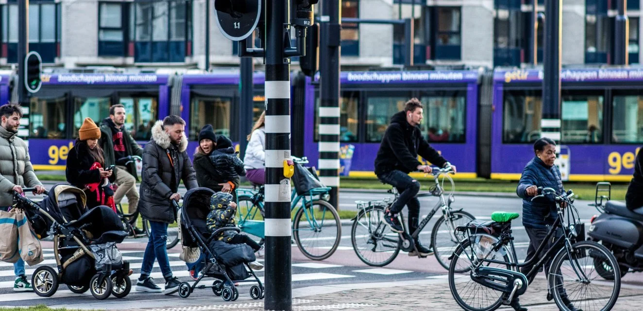 Mensen wachten bij een zebrapad op een stedelijk kruispunt met een trambus en fietspaden, in een druk stadscentrum.