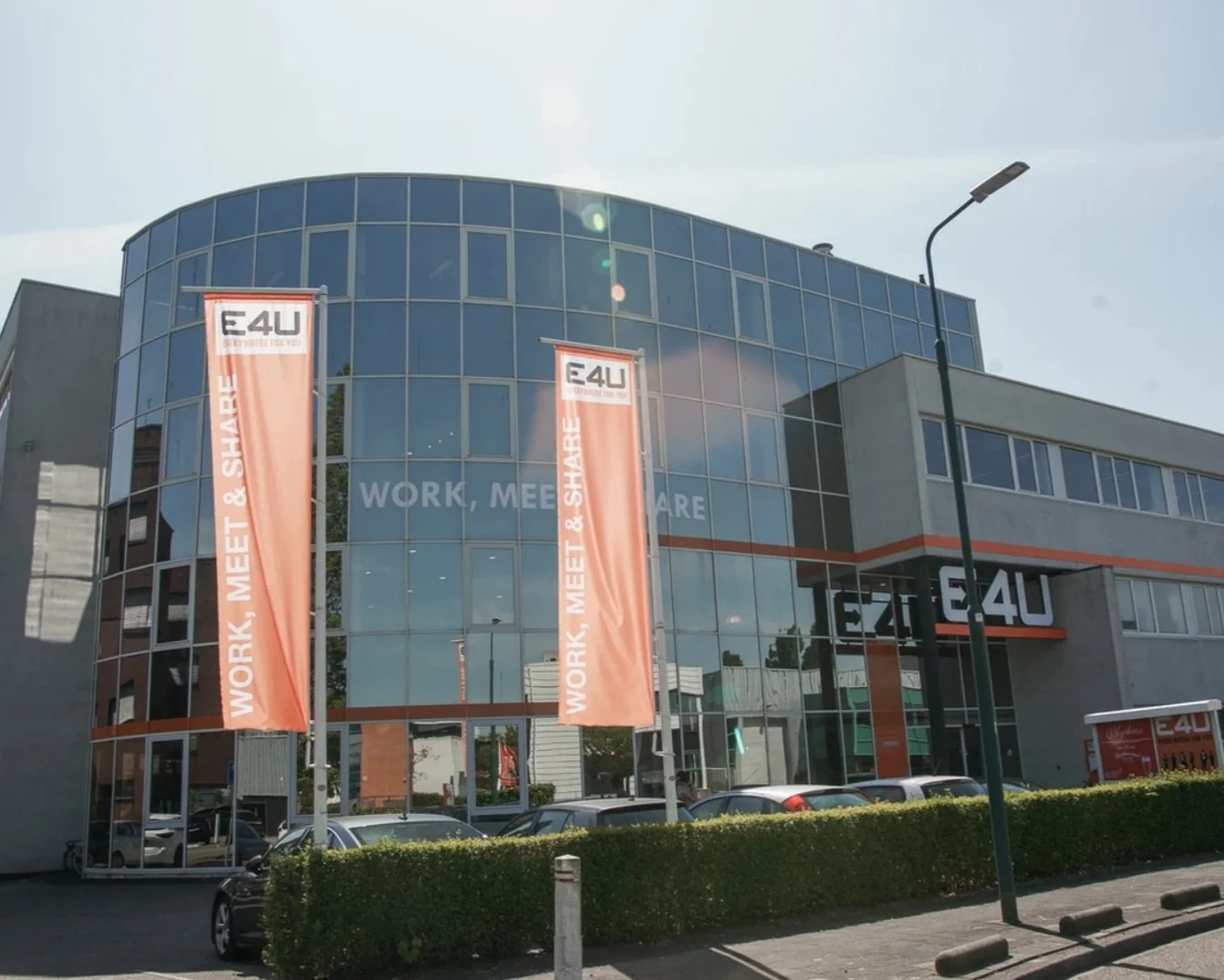 Modern kantoorgebouw met grote glazen gevels en banners die E4U en de slogan 'WORK, MEET & SHARE' tonen, parkeren voor de deur, heldere dag en een lantaarnpaal.
