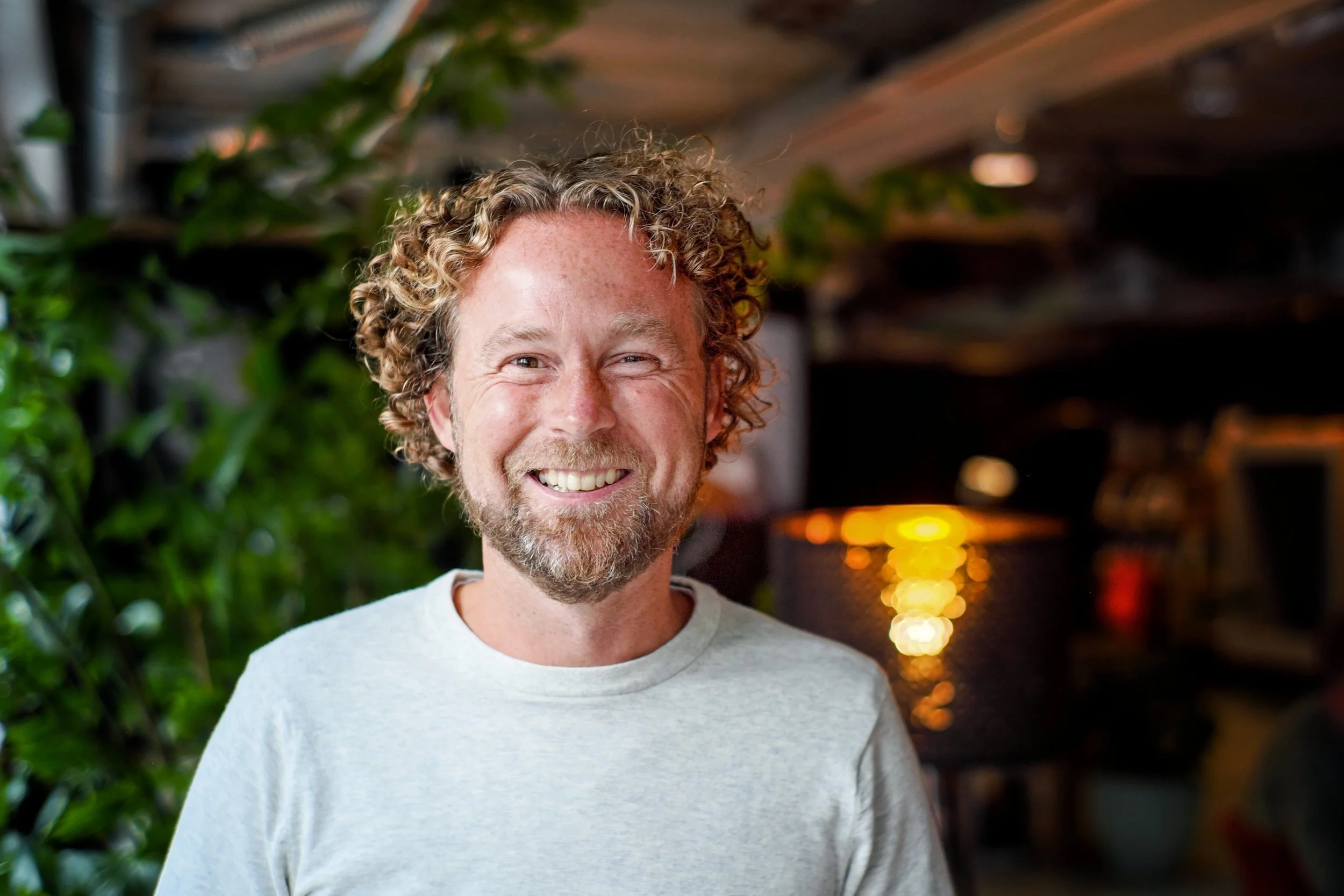 Een lachende man met gekrulde haar en een baard, gekleed in een grijs t-shirt, staat binnen met groene planten en warme verlichting op de achtergrond.