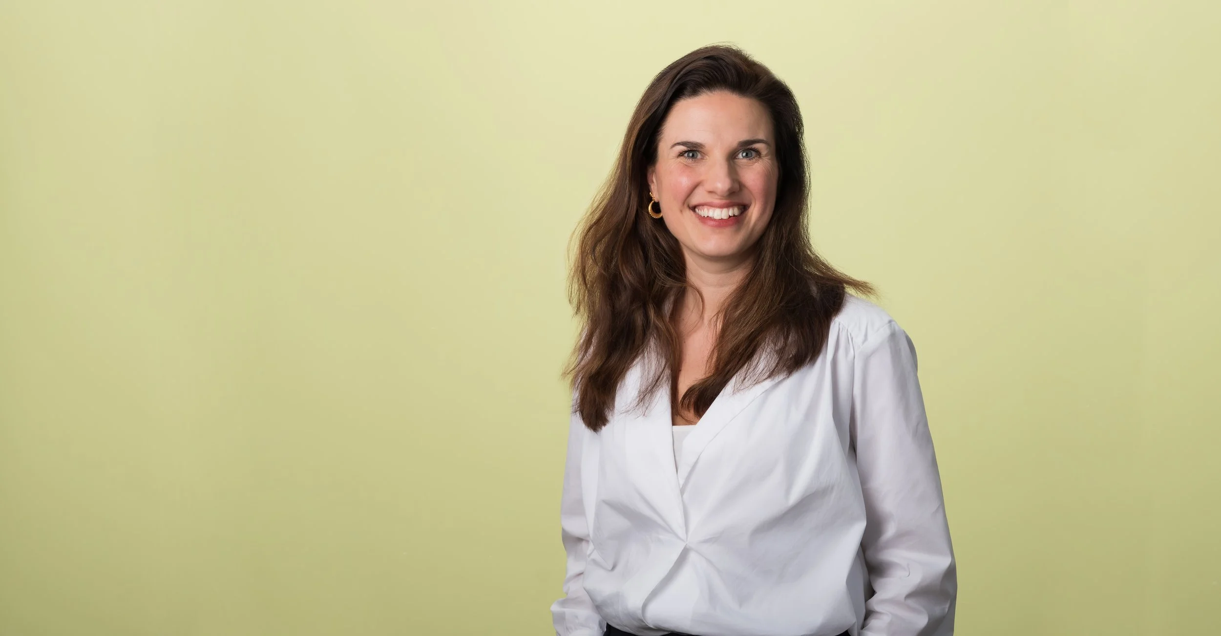 Portret van een lachende vrouw met bruin haar, witte blouse, gouden oorbellen, staand voor een lichte, geelgroene achtergrond.