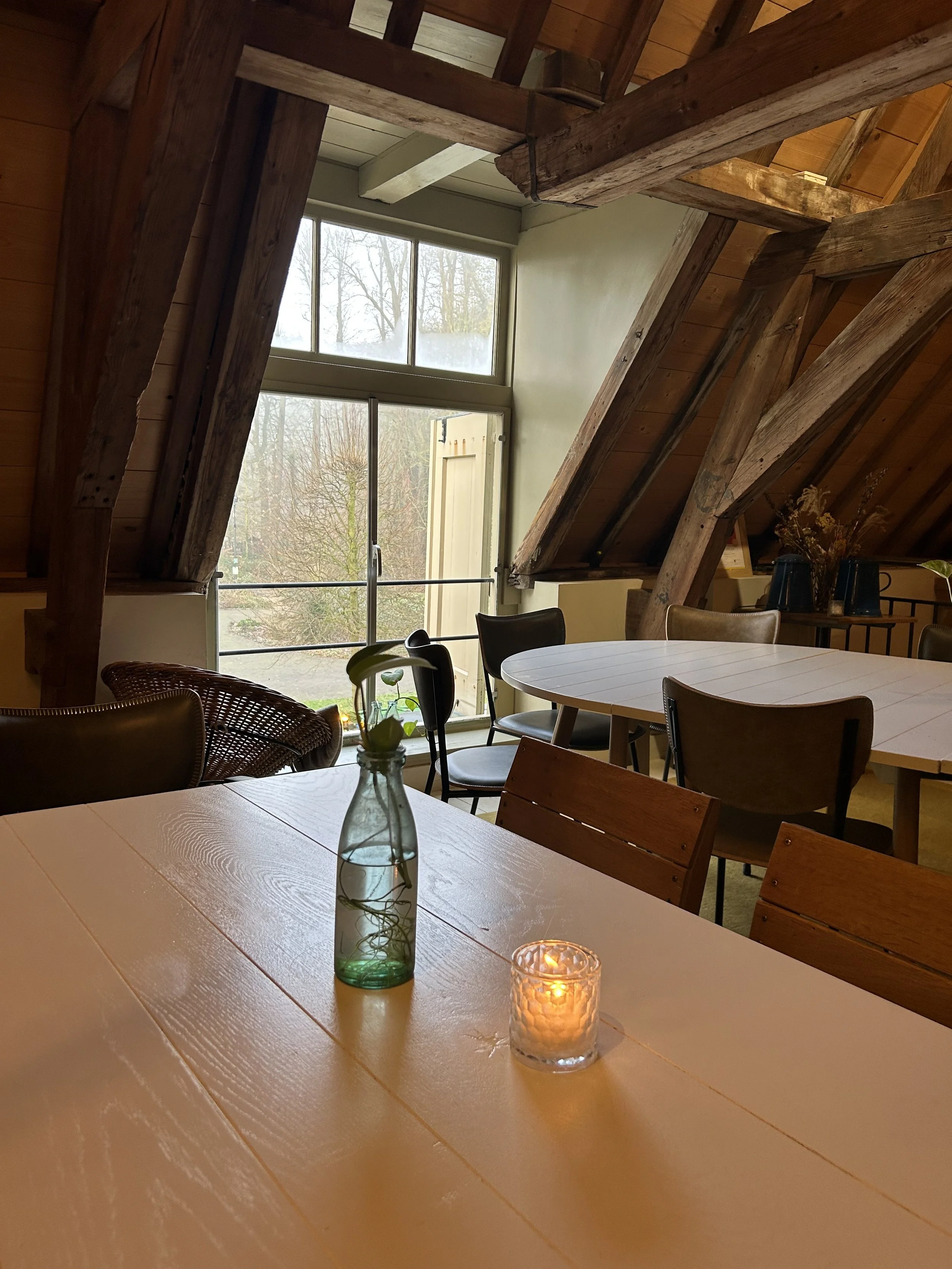 Binnenruimte met tafel, planten, kaarsen en grote ramen met uitzicht op buiten planten en bomen.
