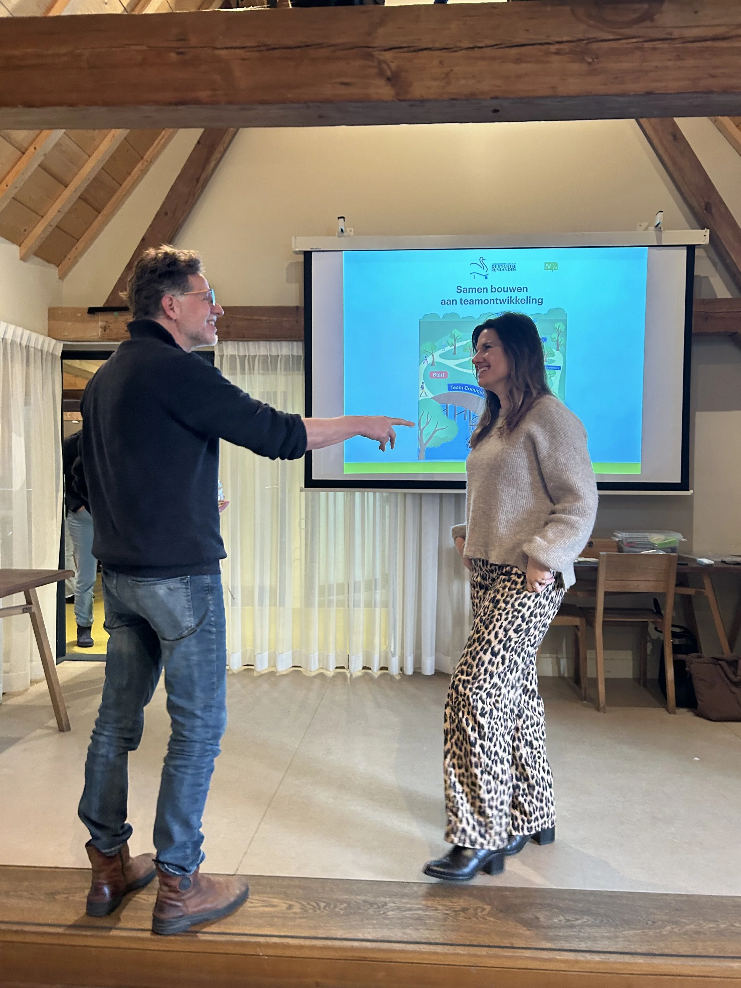 Twee mensen, een man en een vrouw, lachen en praten met elkaar in een kamer met een projector en een scherm aan de muur.