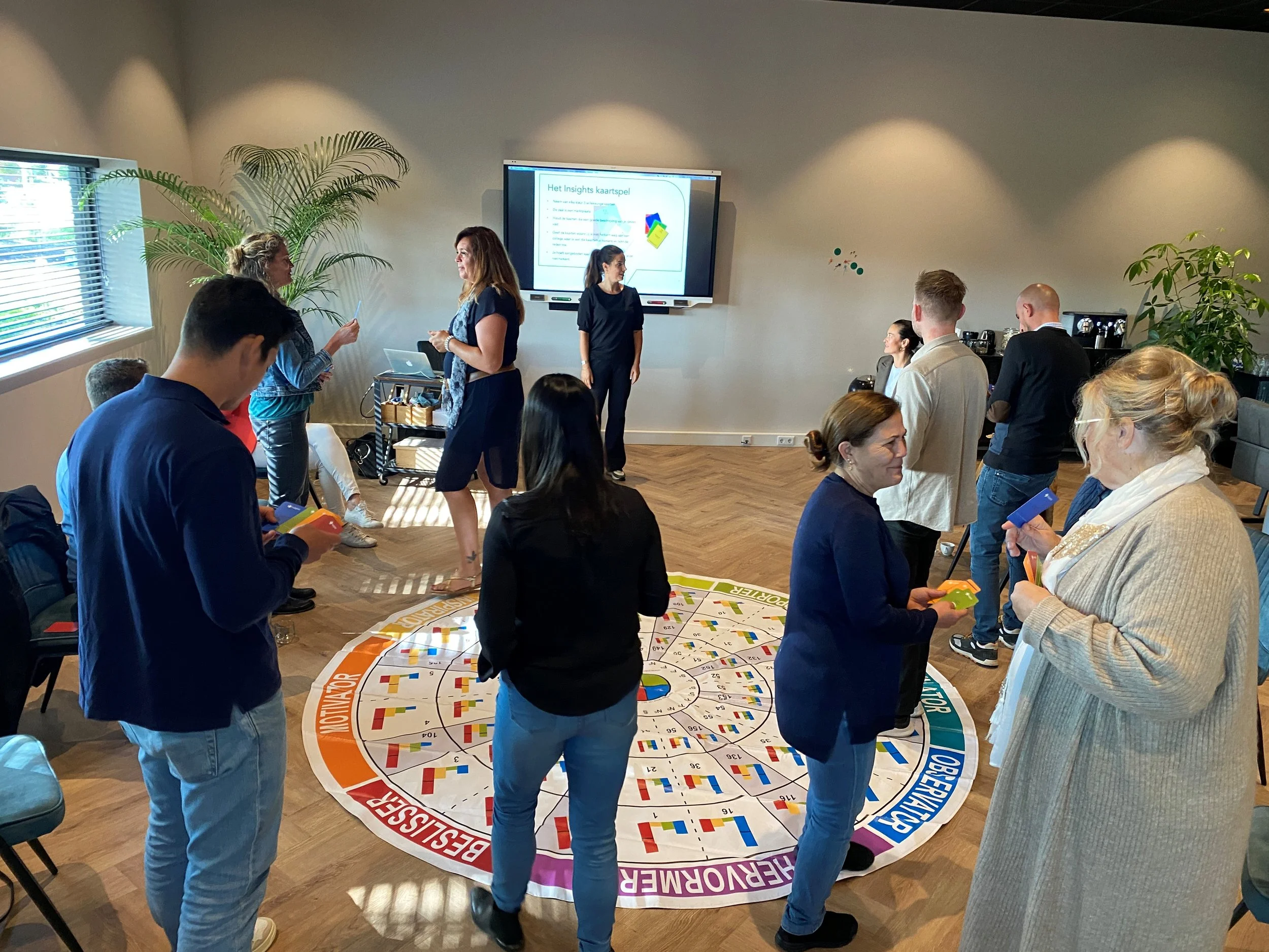 Groep mensen die samen een bordspel spelen in een kamer met planten en een digitale presentatie op een groot scherm.