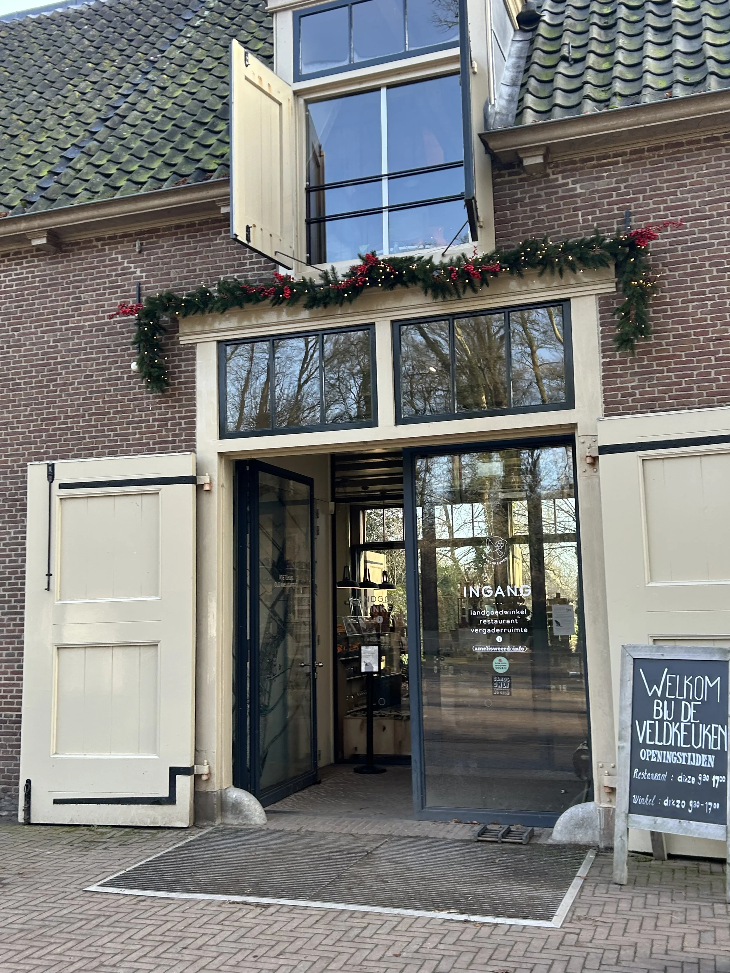 Uitsnede van de ingang van een winkel met een grote glazen deuropening, een zwart uithangbord met witte tekst, en een bord met de tekst 'WELKOM BIJ DE VELDKUEKEN'. Boven de ingang hangt een groene kerstversiering met rode bessen en kleine lichtjes. Het gebouw heeft een bakstenen gevel en een dak met groene dakpannen.