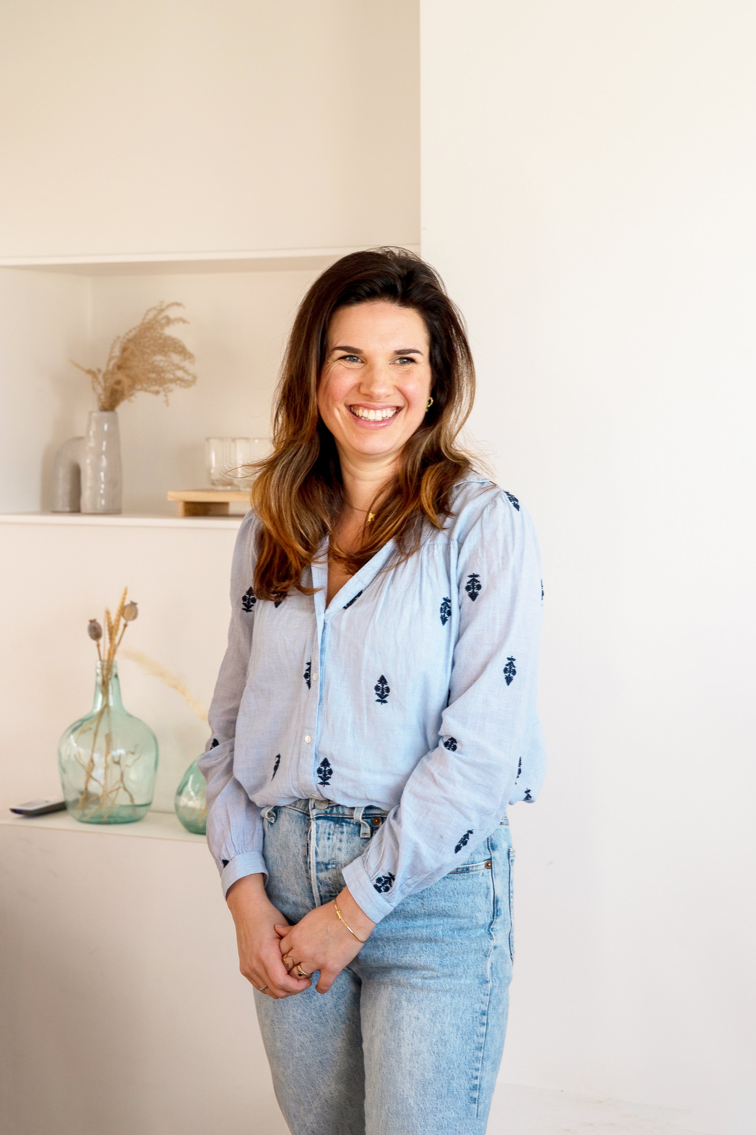 Een glimlachende vrouw met lang bruin haar, gekleed in een lichtblauw overhemd met donkere patronen en lichtblauwe jeans, staat in een minimalistische kamer met witte muren. Op de achtergrond staan decoratieve vazen en glazen.