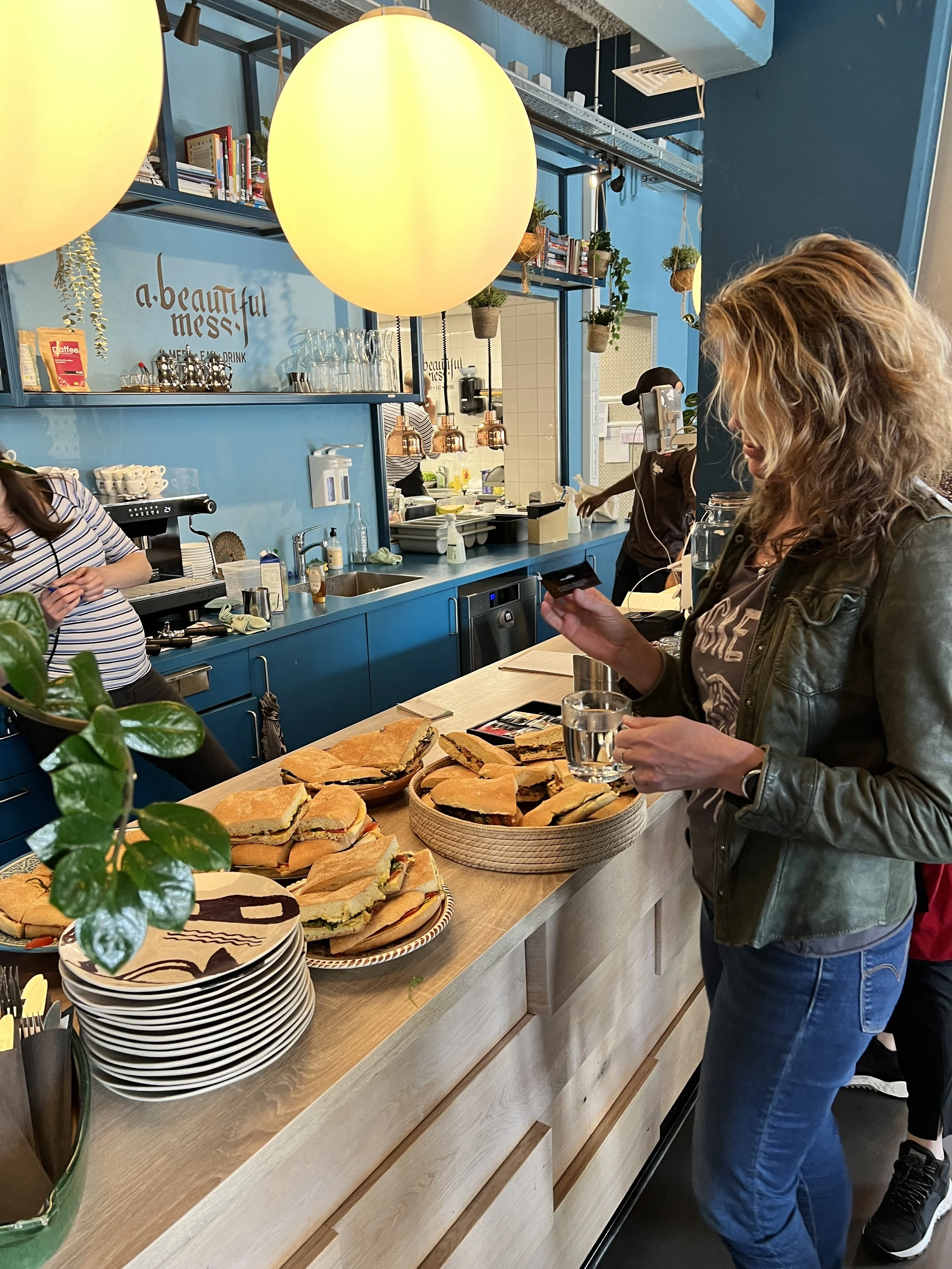 Een vrouw die een glas vasthoudt en een tafel met sandwiches en serveermokken in een café of winkel.