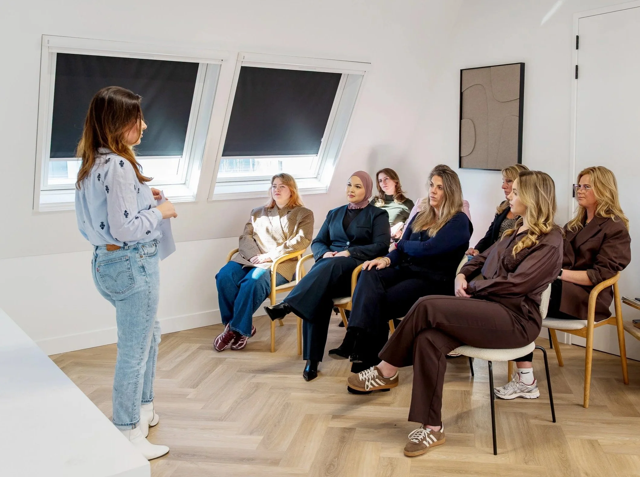 Vrouw staat voor een kleine groep vrouwen die aandachtig luisteren. De setting lijkt een informele bijeenkomst of workshop, met een minimalistisch interieur en natuurlijke lichtinval door grote ramen met verduisteringsgordijnen.