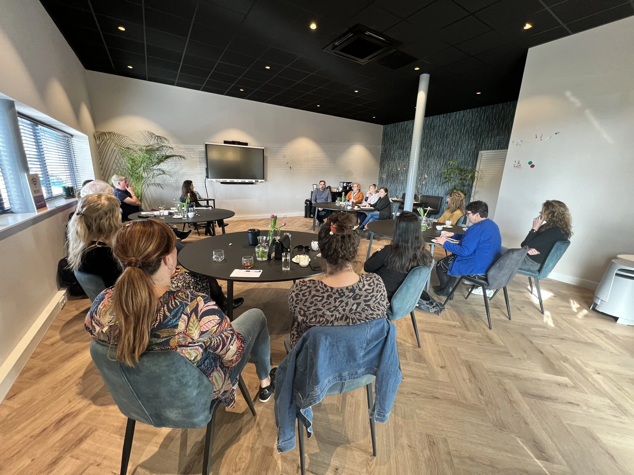 Groep mensen zit rond een tafel in een vergaderzaal, terwijl een man en een vrouw aan het praten zijn. De zaal heeft een groot scherm en decoraties, planten en ramen met jaloezieën.