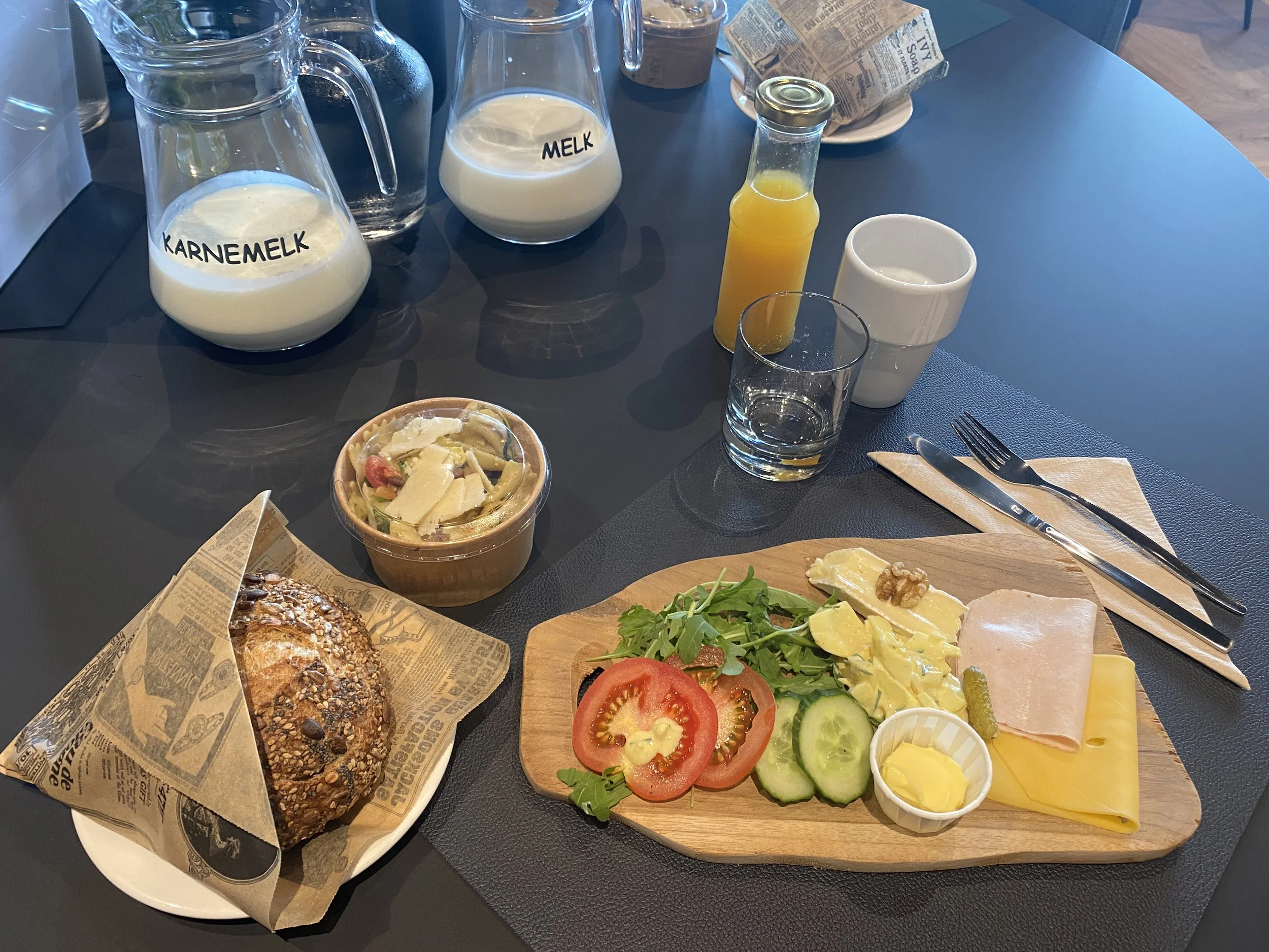 Een tafel met diverse herinneringsproducten voor een baby, waaronder jampotten met melk en karnemelk, een glas sinaasappelsap, een schaal met salade, een stuk brood met haver en zaden, en een bord met gesneden tomaten, komkommers, sla, en kaas.