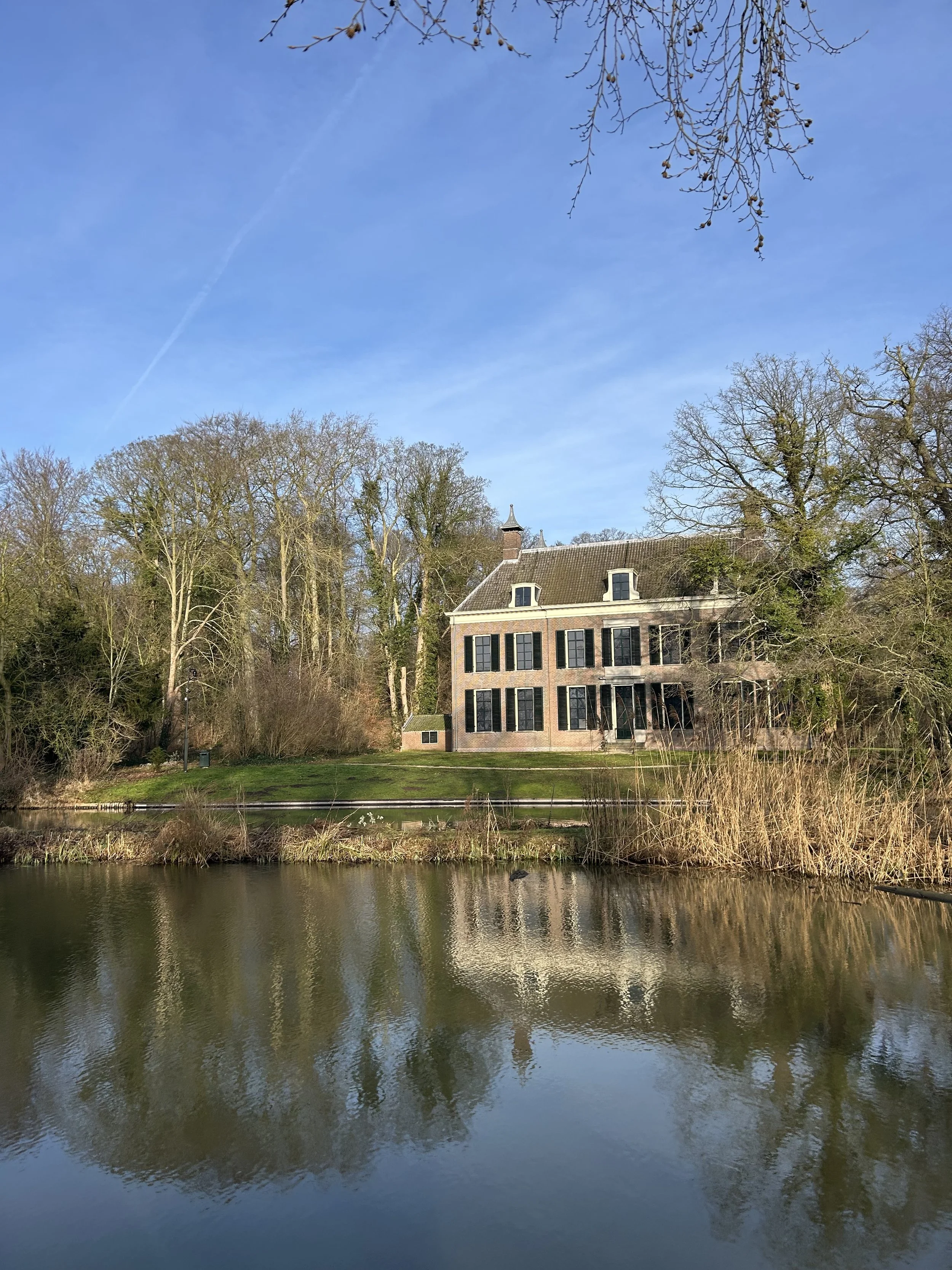 Een gebouw met zwarte raamkozijnen aan de rand van een waterkant, omgeven door bomen zonder bladeren en een heldere blauwe lucht.