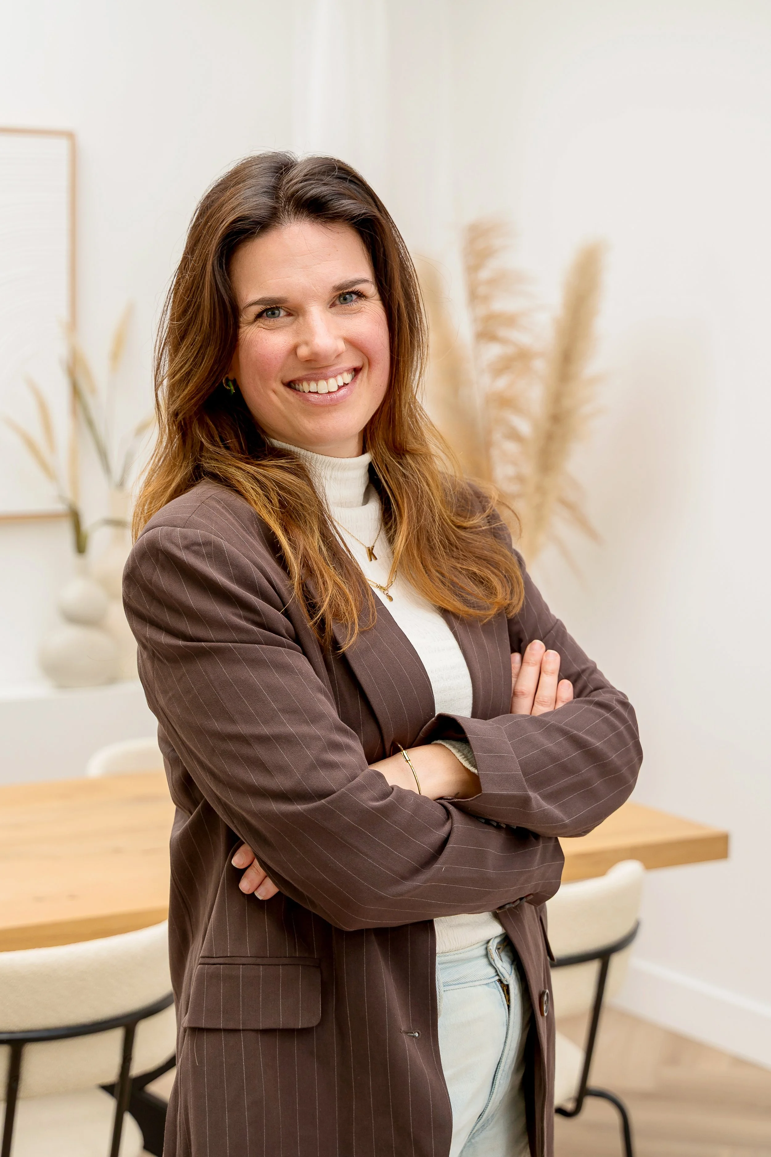 Een vrouw met bruin haar draagt een bruine blazer en witte sweater, lacht en staat met gekruiste armen in een vergaderruimte.