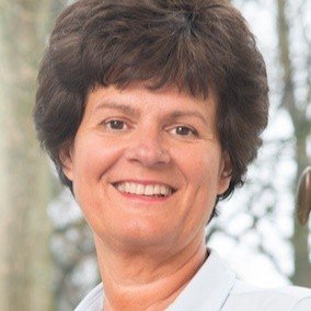 Portret van een lachende vrouw met korte, krullende bruinhaar, achtergrond van bomen en natuurlijk licht.