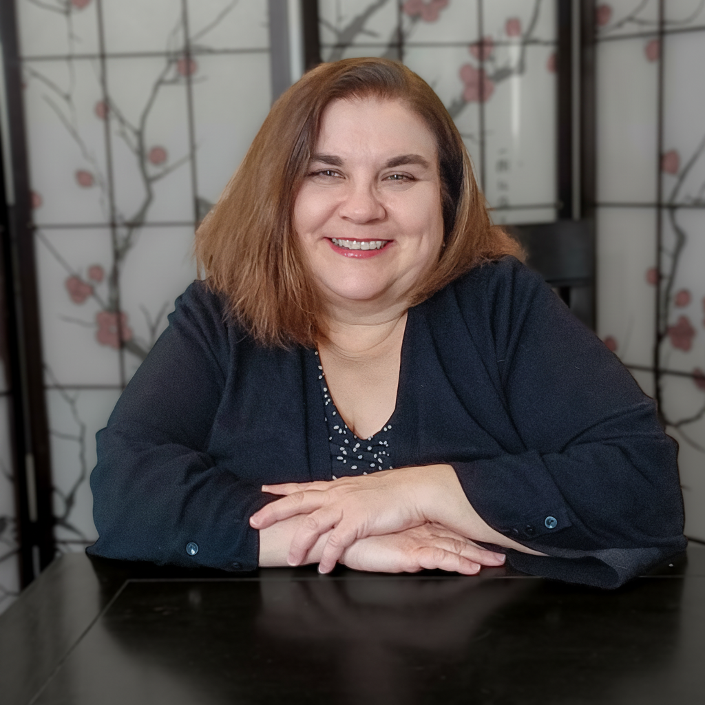 A woman with shoulder-length light brown hair smiling and sitting at a black table, with a decorative background featuring floral patterns.