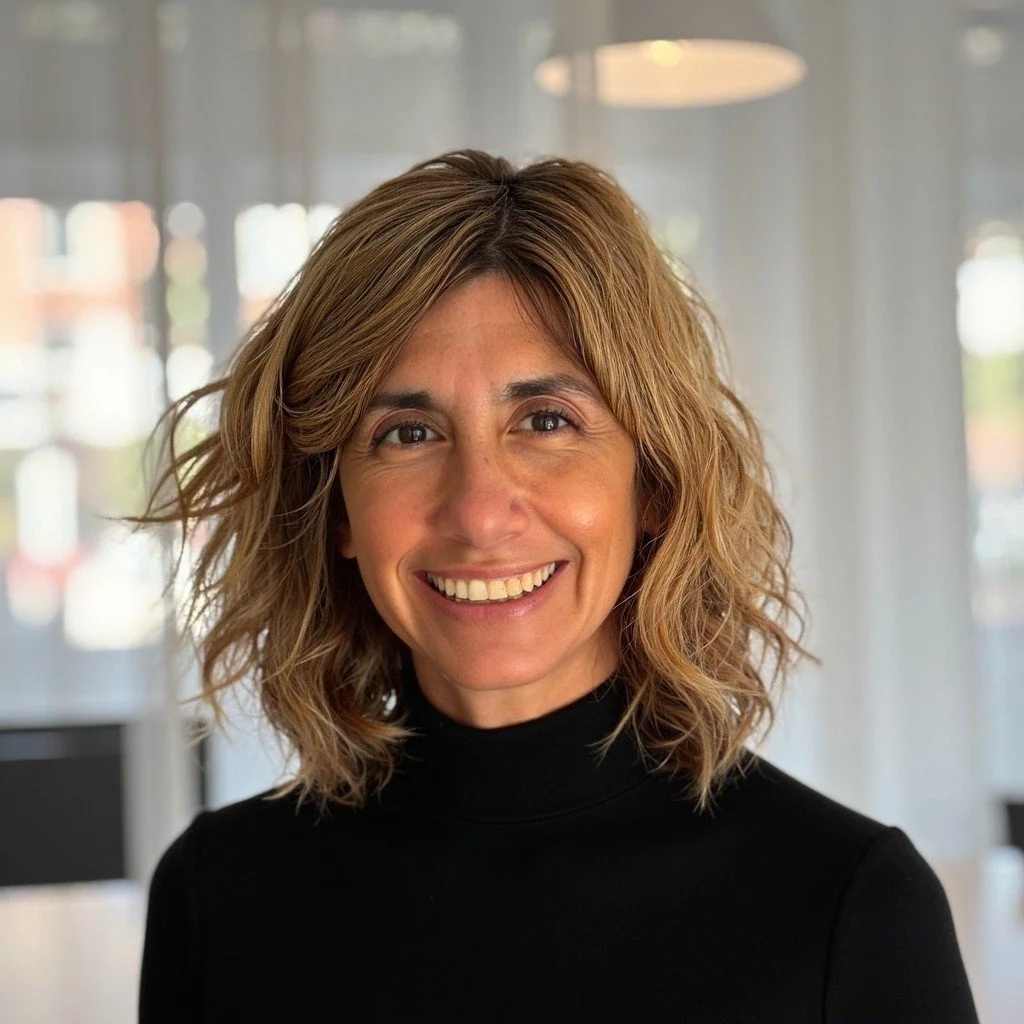 A smiling woman with wavy, shoulder-length hair wearing a black turtleneck in an indoor setting with blurred windows and lights in the background.