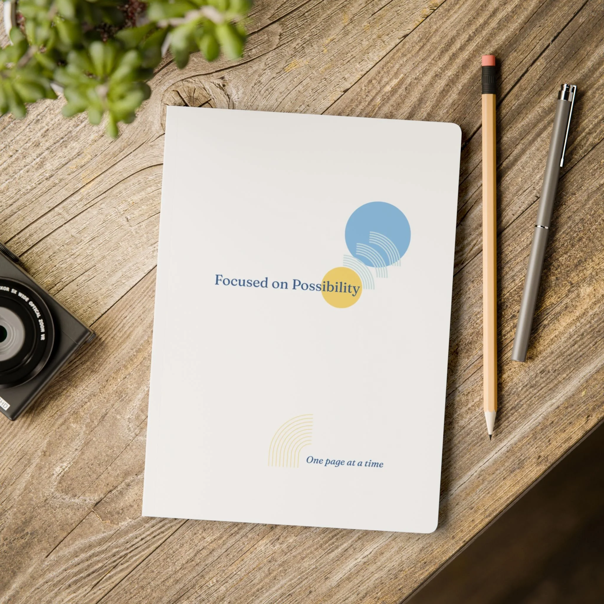 A wooden desk with a camera, a notebook titled 'Focused on Possibility', a pink-and-black pen, and a silver pen. The notebook has circles and the phrases 'One page at a time' and 'Focused on Possibility' on the cover.