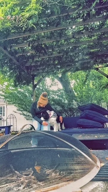Person in a beanie and jacket petting a small white dog in an outdoor area with trees and covered roof, surrounded by stacked luggage and a car.