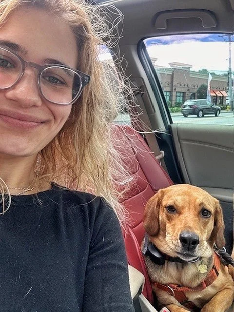 A woman with curly blonde hair, glasses, and a black shirt taking a selfie inside a car, with a brown dog sitting in the backseat next to her, wearing a collar and harness.