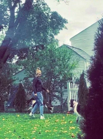 A woman walking with three dogs in a backyard with green grass, trees, and a white picket fence.