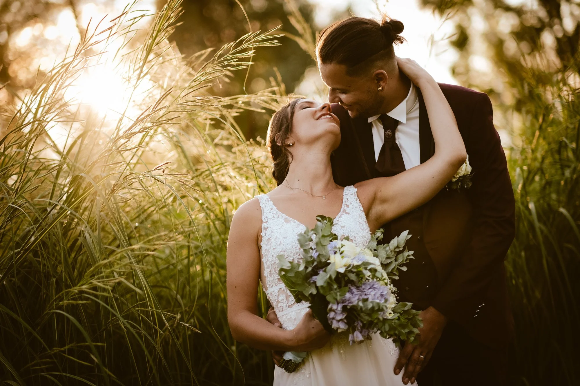 Un couple en robe de mariage et costume se tient dans un champ avec des herbes hautes au coucher du soleil, partageant un moment d'intimité et de tendresse.