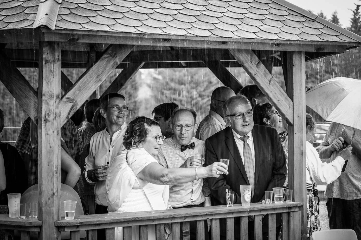Groupe de personnes souriantes et discutant sous une structure en bois lors d'une fête ou d'un événement en plein air, en train de porter un toast, avec des boissons dans les mains.