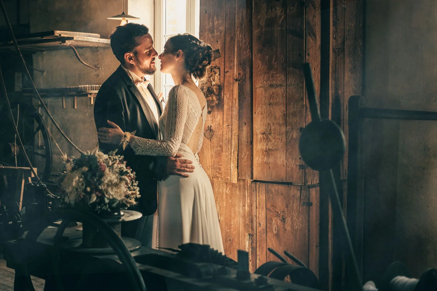 Un couple en habits de mariage, s'embrassant dans une pièce rustique éclairée par la lumière naturelle venant d'une fenêtre.