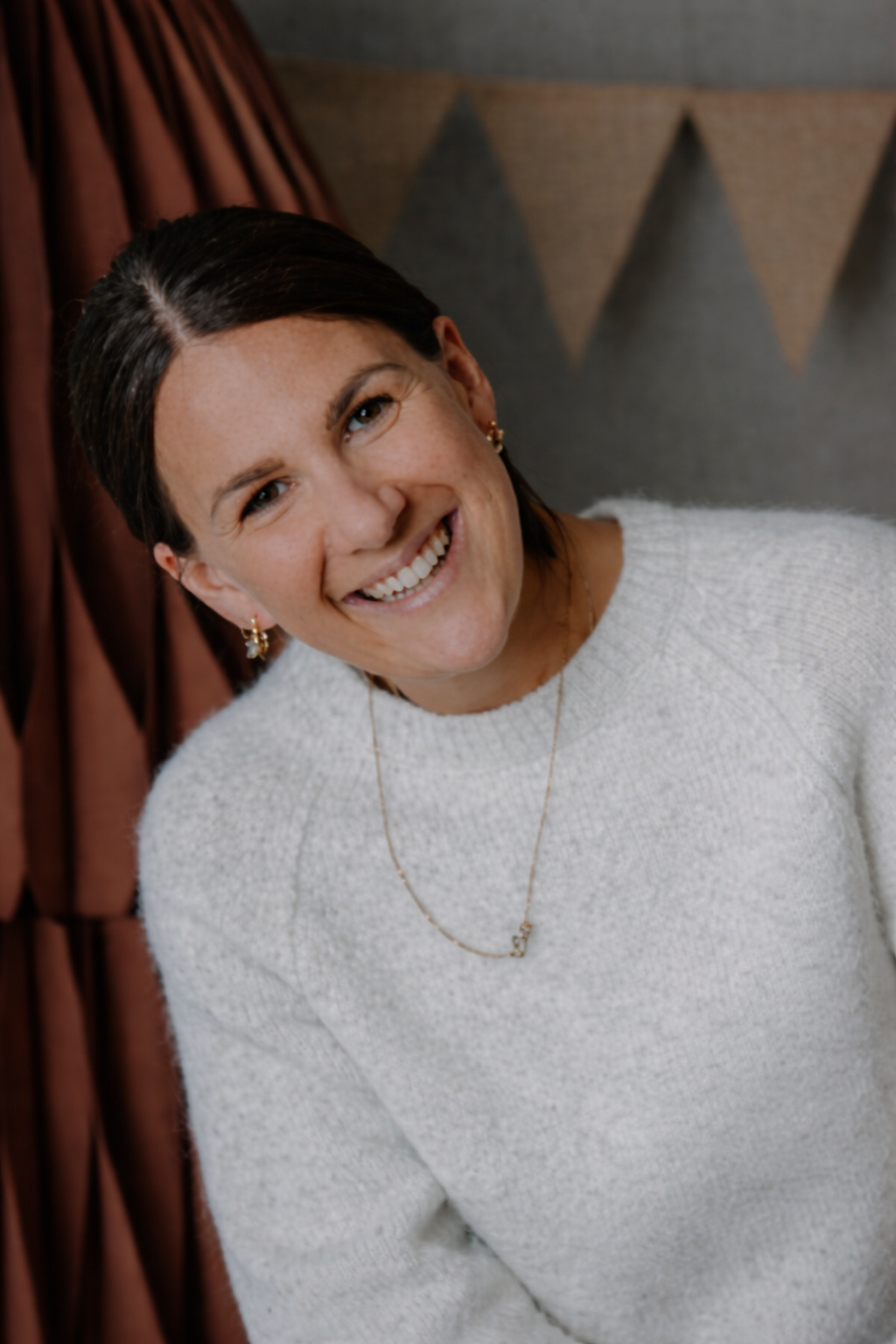 Lächelnde Frau mit dunklem Haar, weiße Pullover, Goldschmuck, vor buntem Hintergrund.