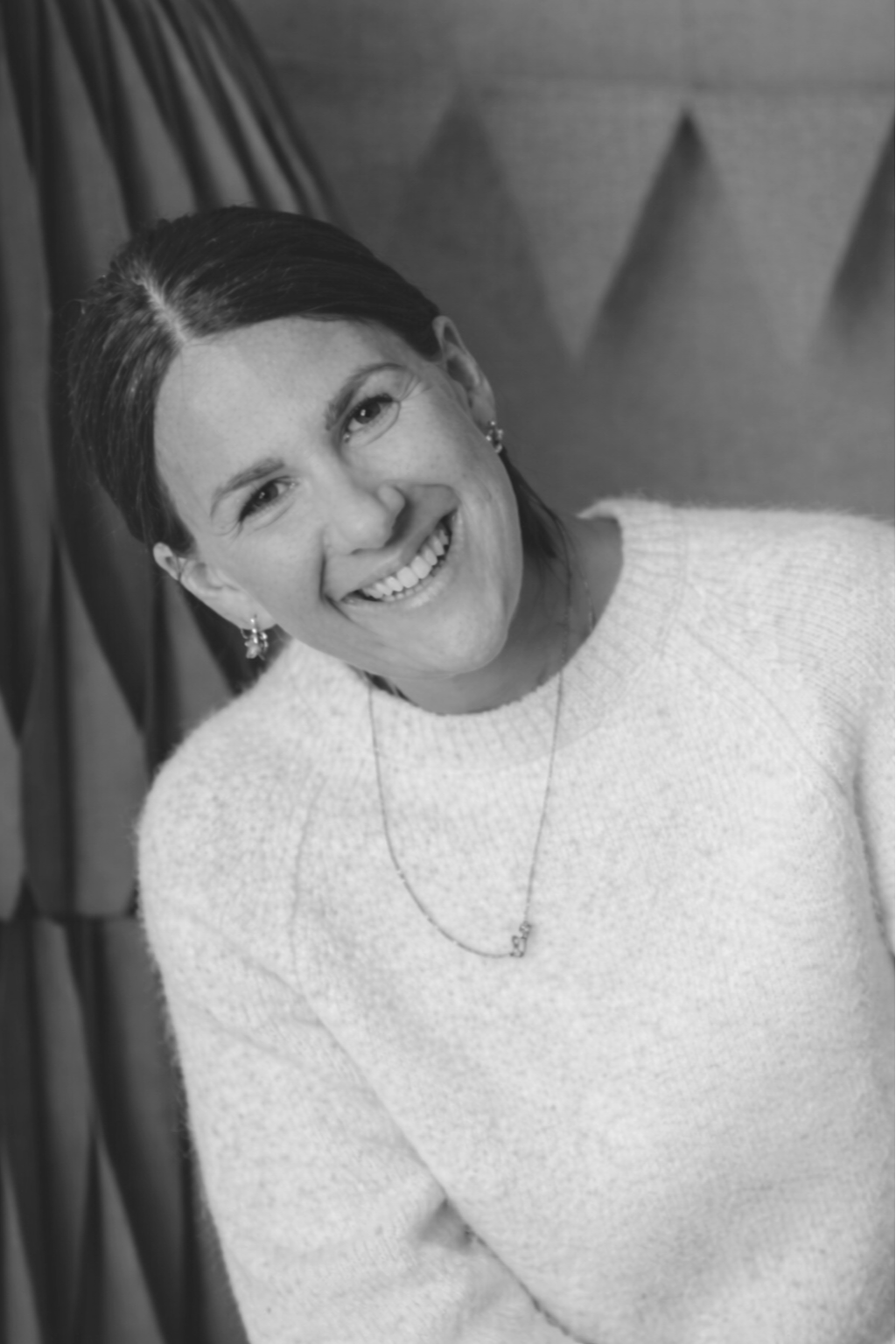 Eine lächelnde Frau mit dunklem, glattem Haar, die ein cremefarbenes Pullover trägt, vor einem Hintergrund mit Vorhängen.