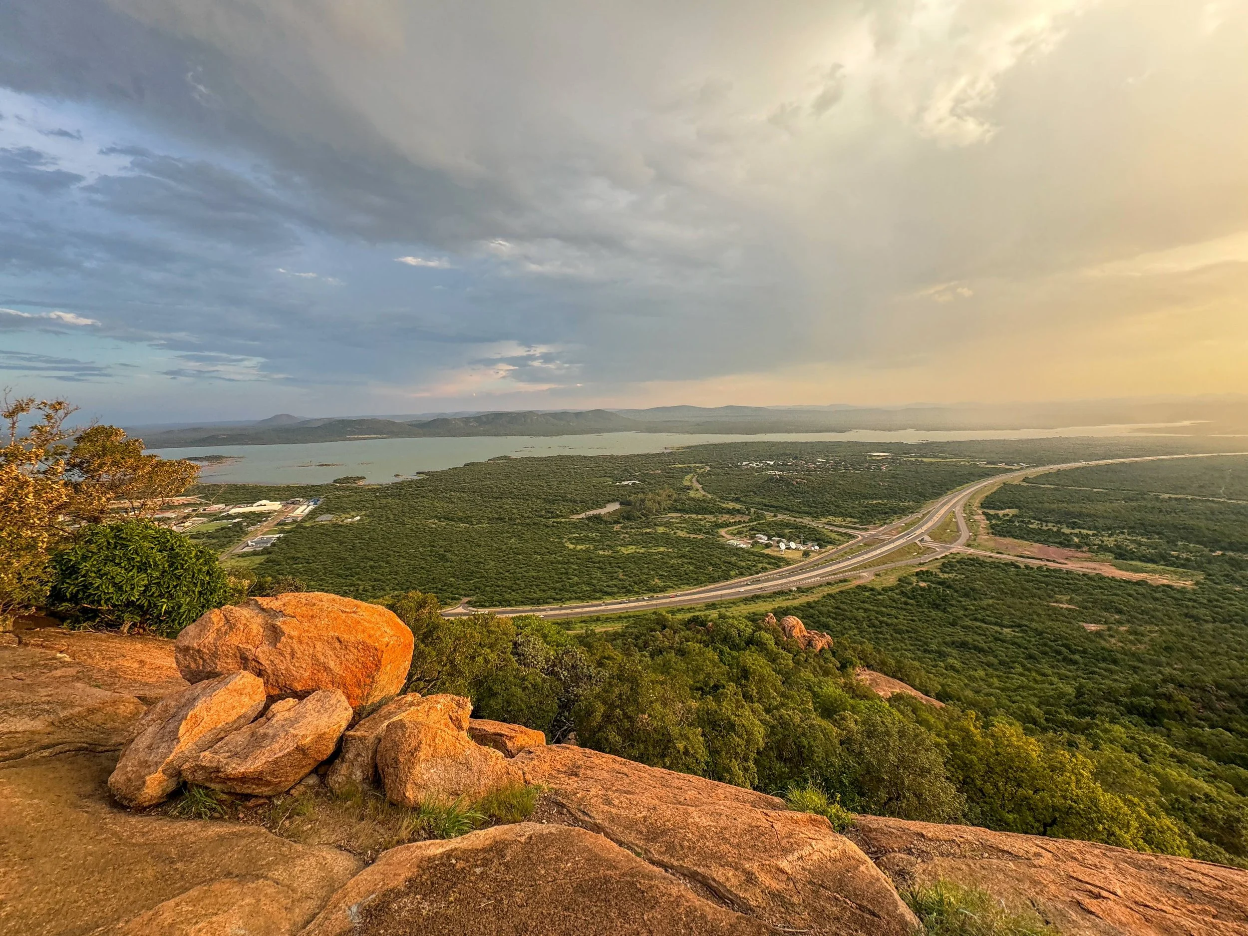 A scenic view from a high vantage point showing a landscape with green vegetation, a body of water in the background, a road curving through the landscape, and a rocky foreground with some trees.